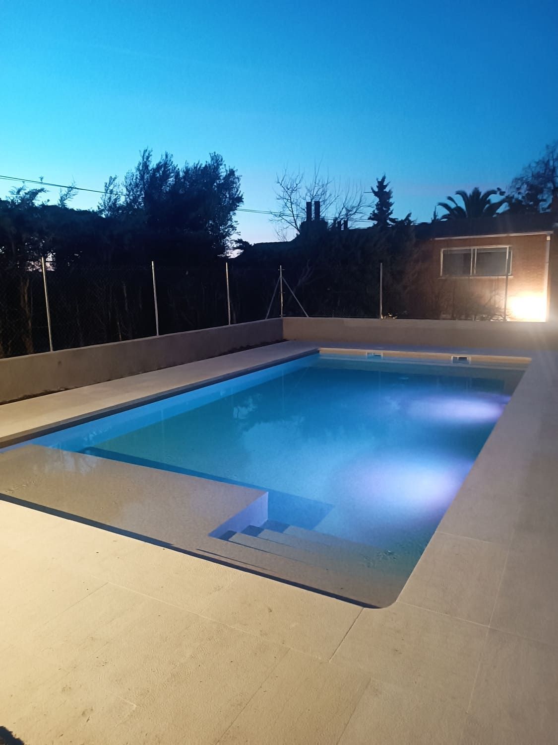 Piscina iluminada al anochecer, con escaleras que conducen al agua. Patio y jardín trasero circundantes.