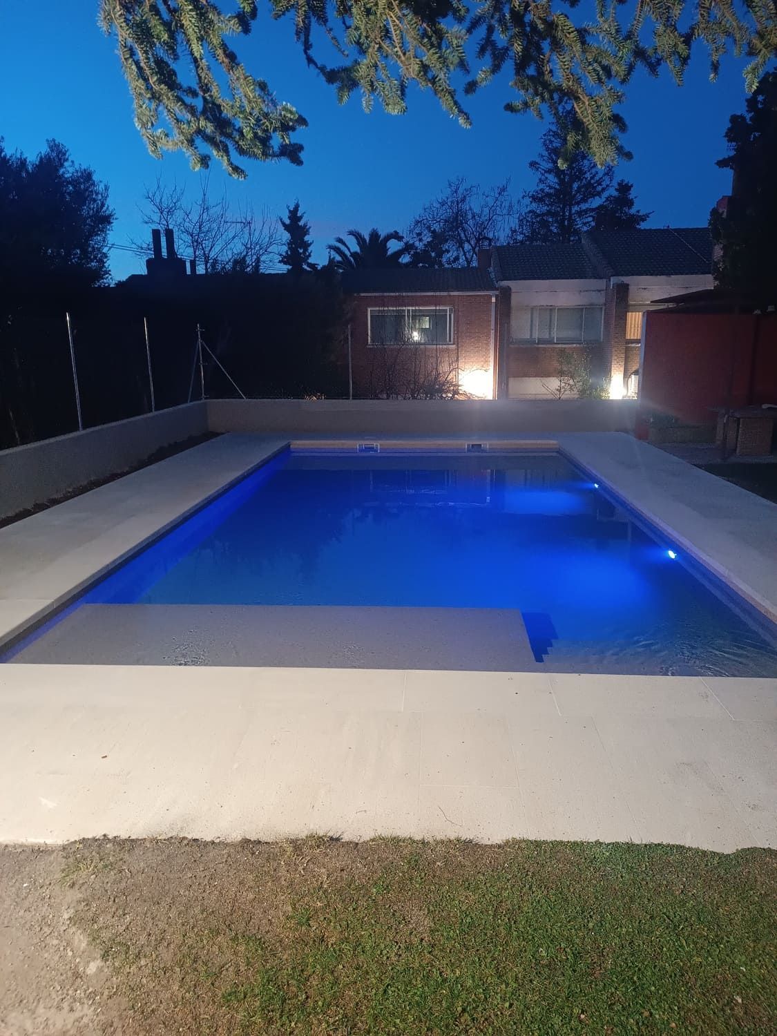 Piscina iluminada de noche con luces azules, rodeada de un borde de hormigón y una casa al fondo.