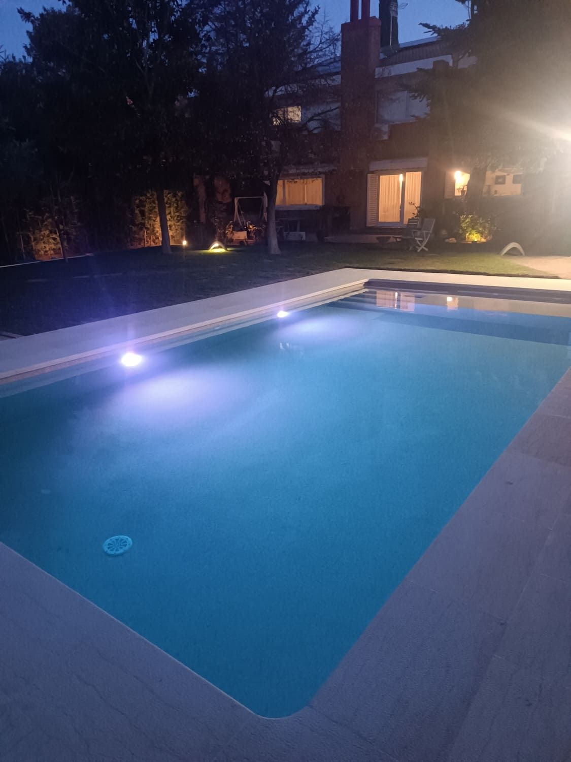 Vista nocturna de una piscina rectangular con luces subacuáticas, con vista a una casa con luces.
