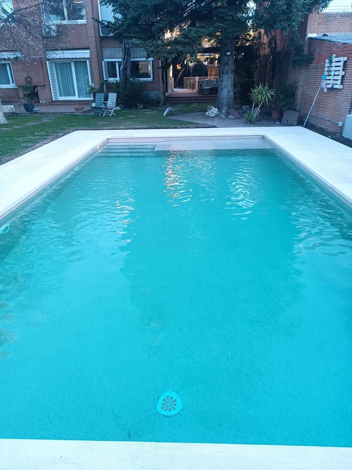 Piscina de agua azul cristalina con borde de hormigón, ubicada en un patio trasero. Edificios al fondo.