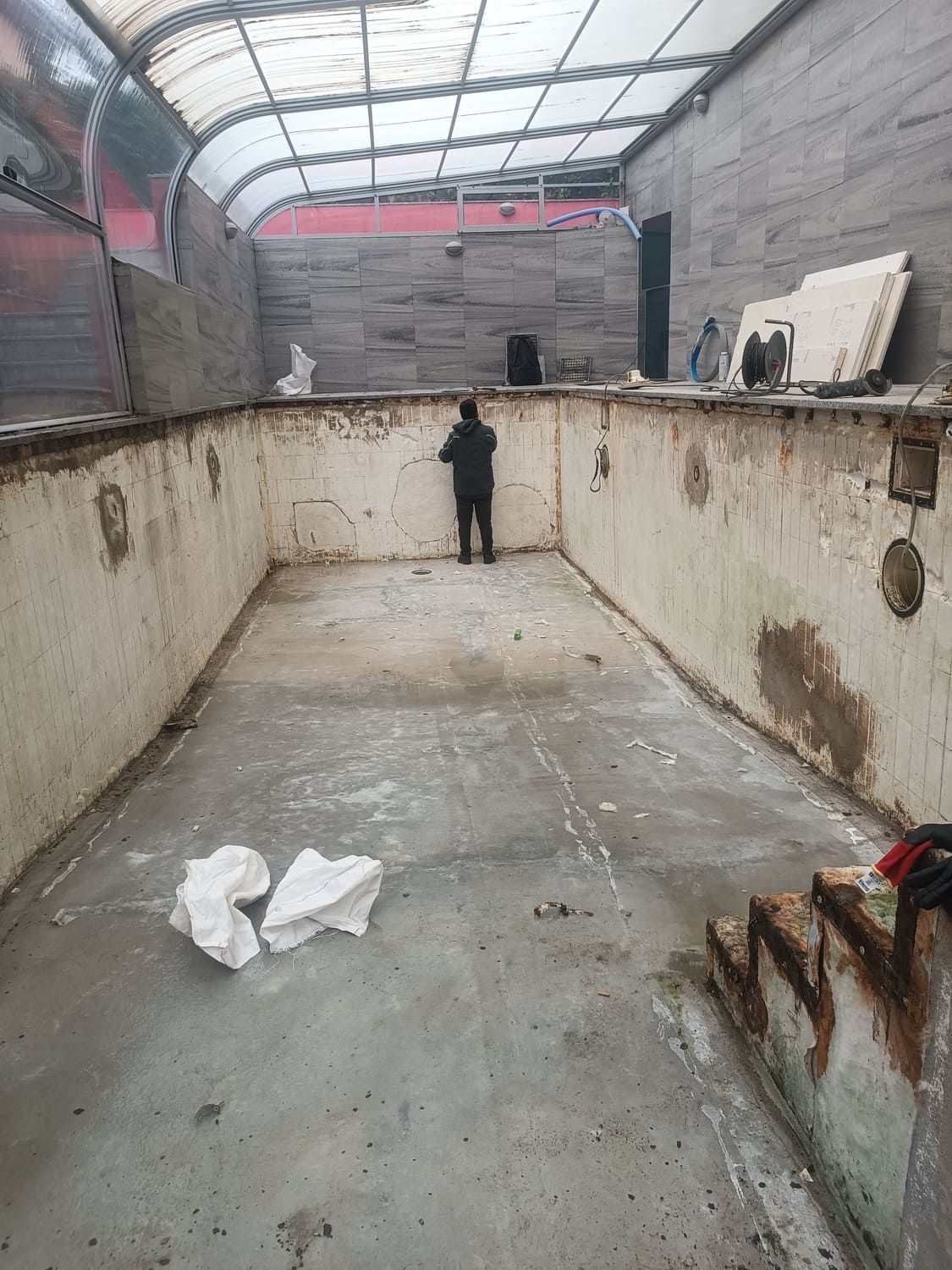 Piscina cubierta vacía, con una persona dentro. Las paredes y el suelo de la piscina son de hormigón, con escombros esparcidos.