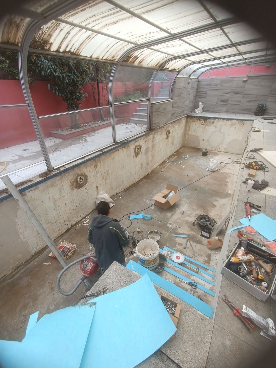 Persona trabajando dentro de una piscina vacía debajo de una estructura parcialmente cubierta; herramientas y escombros visibles.