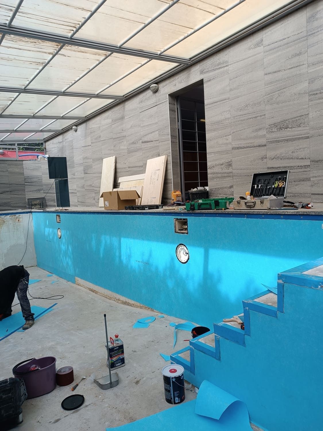 Piscina en construcción; interior pintado de azul, con herramientas y materiales presentes. Persona trabajando cerca del borde de la piscina.