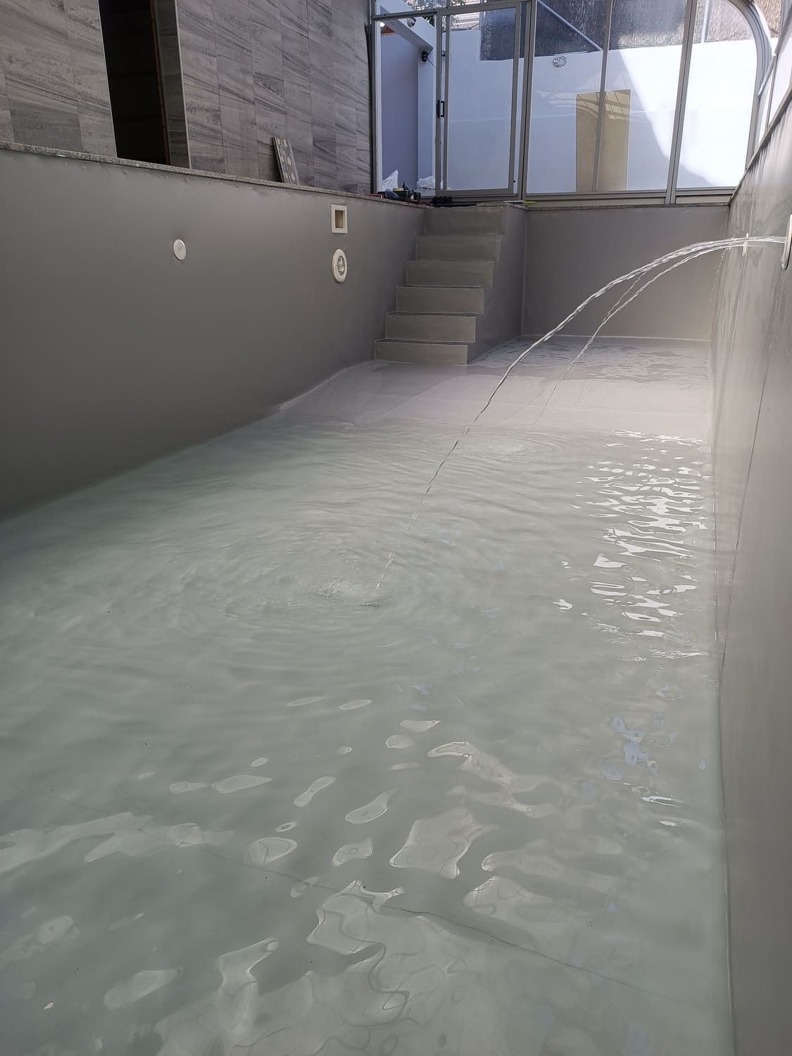 Piscina cubierta llena de agua turbia. Paredes grises, escalones y fuente.