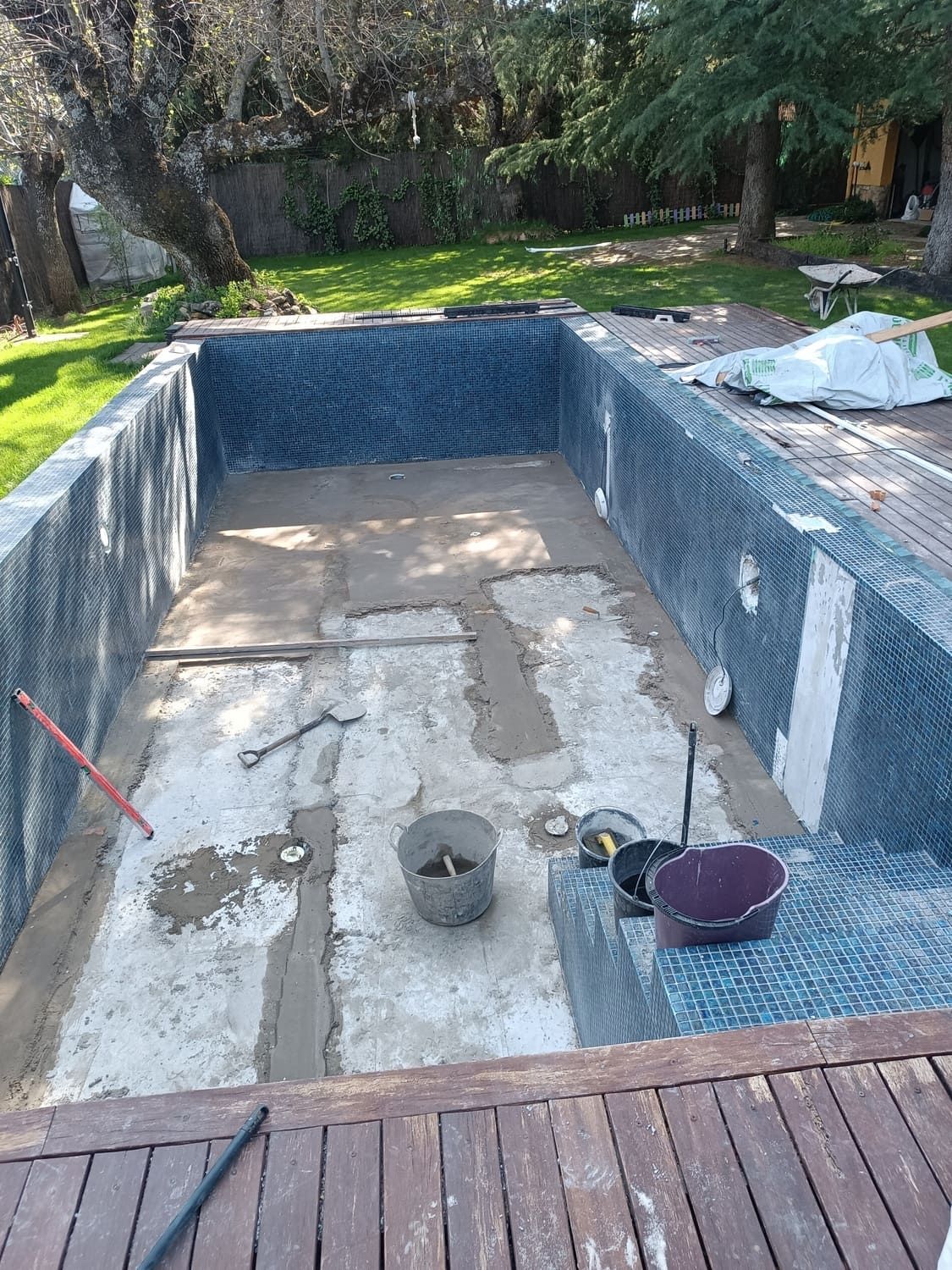 Piscina en construcción; baldosas azules, interior de hormigón, rodeada de tarima de madera.
