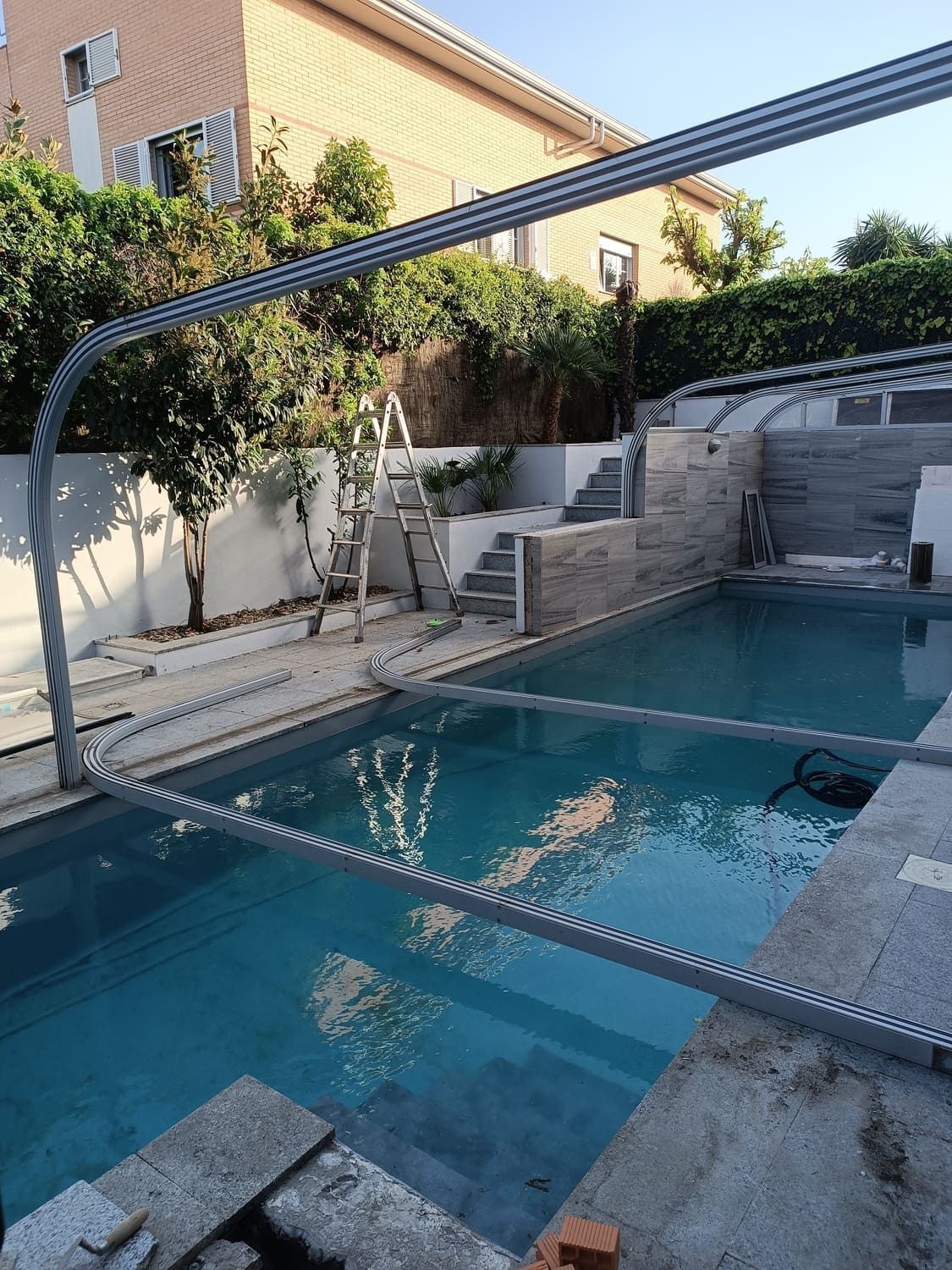 Piscina en construcción; estructura de metal en lo alto; escalera cercana; edificio de ladrillo al fondo.