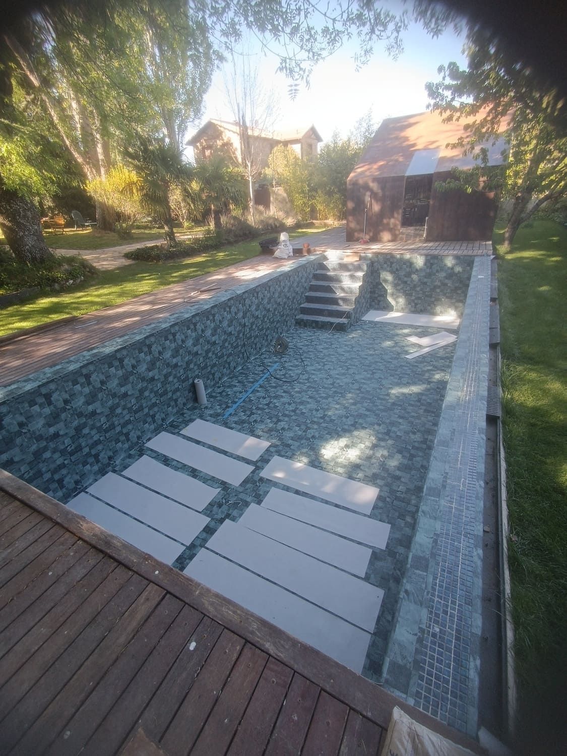 Una piscina rectangular con escalones grises y losas rodeada por una plataforma de madera y azulejos verdes, en un entorno de patio trasero.