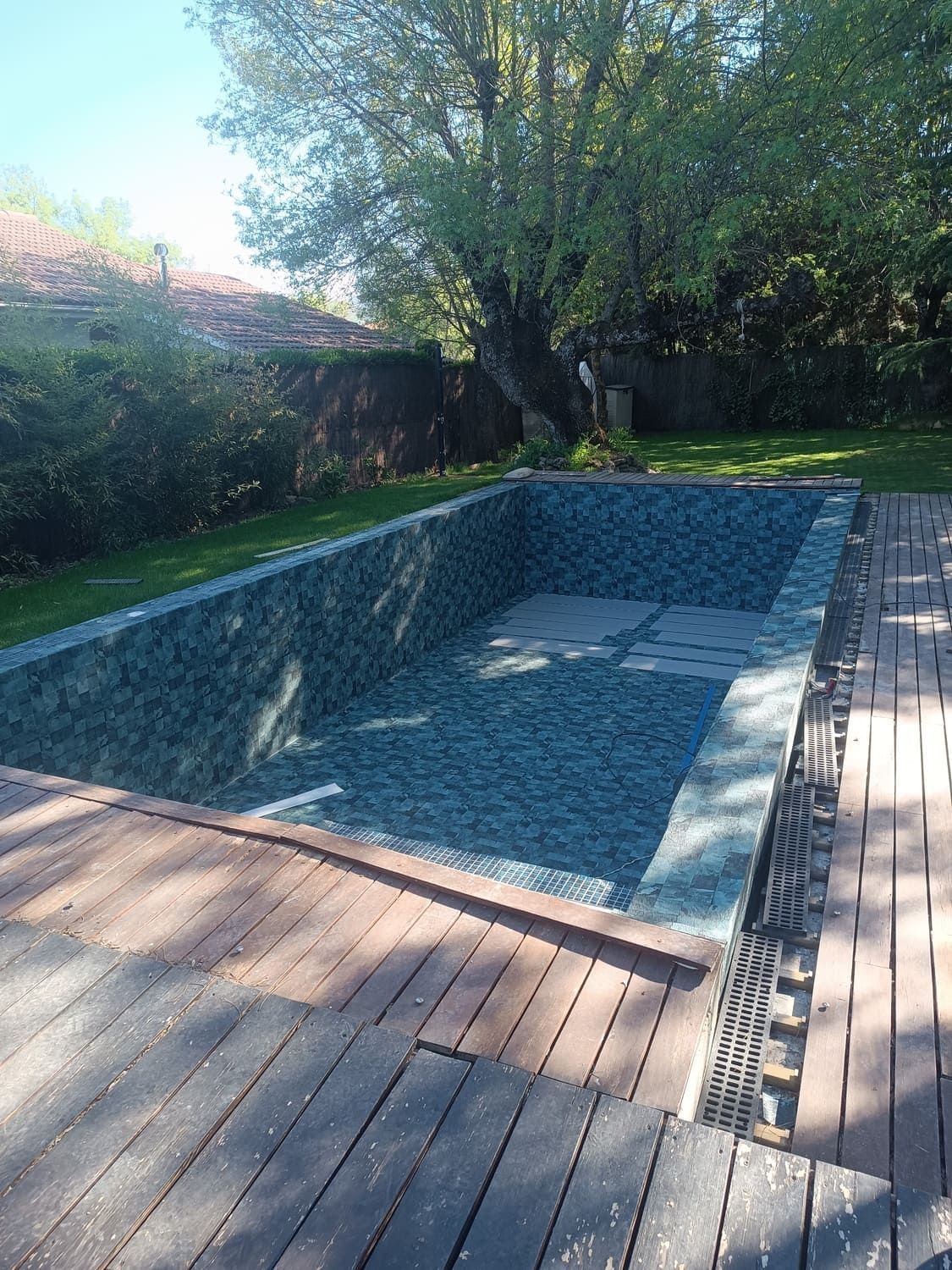 Piscina rectangular vacía con paredes de azulejos verdes, rodeada de tarima de madera y césped.