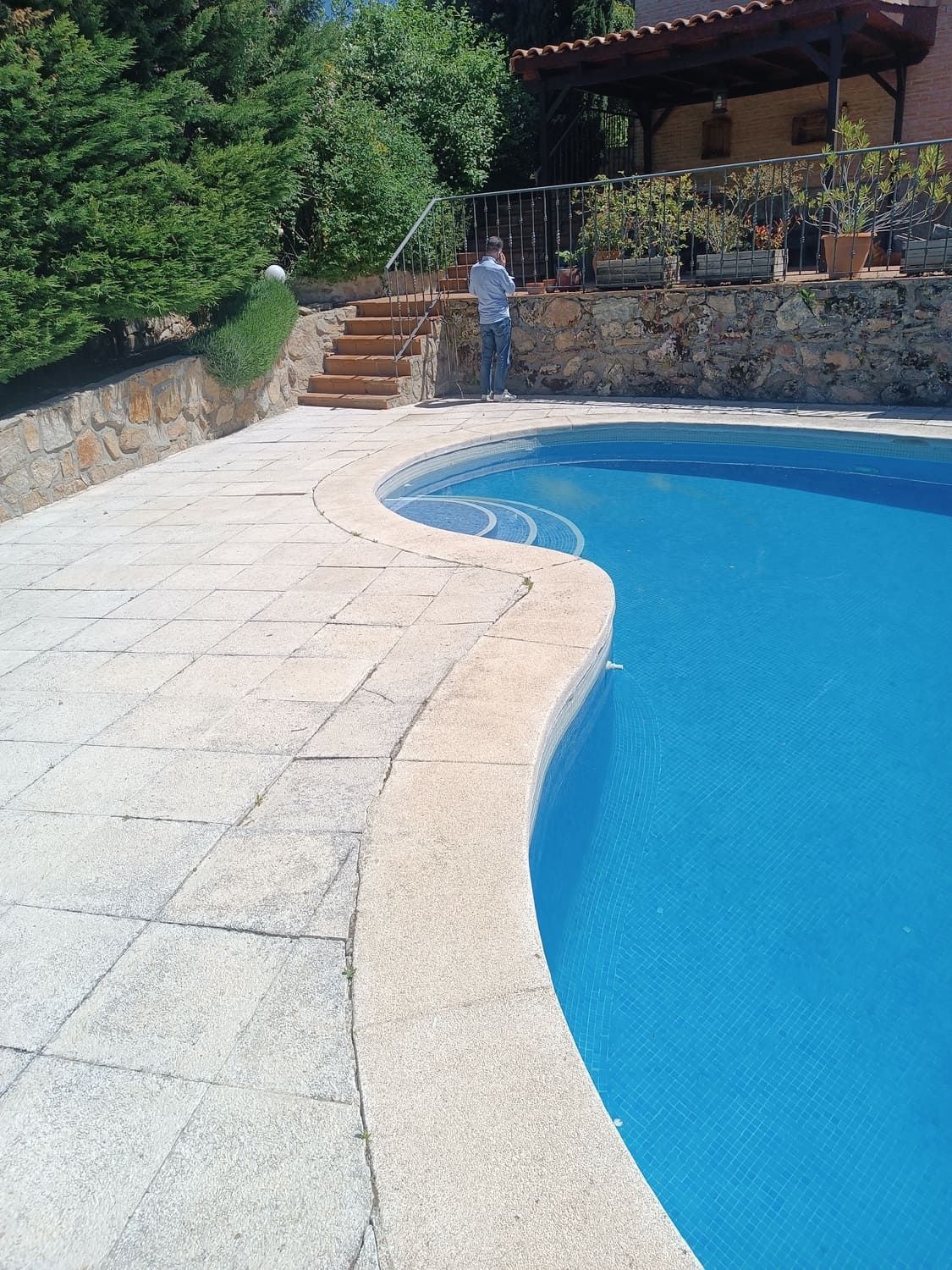 Piscina con agua azul y patio de piedra. Persona de pie junto a las escaleras que conducen a un edificio.