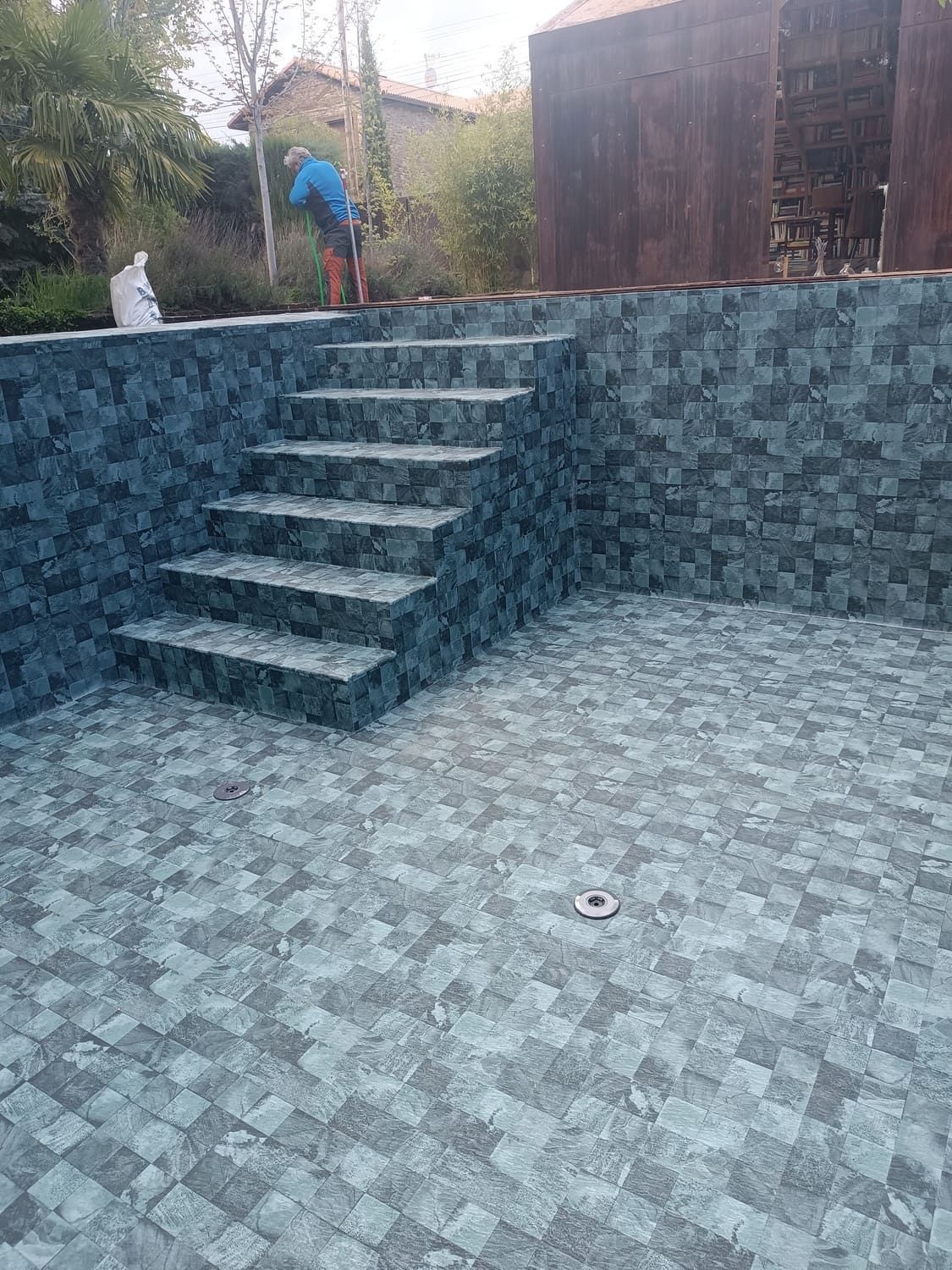 Piscina con escalones y paredes de azulejos azules. Persona trabajando cerca del cobertizo. Exterior soleado.