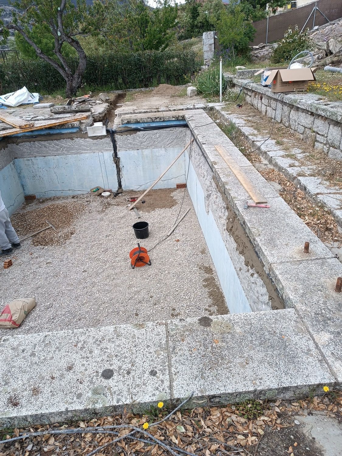 Piscina en construcción, estructura de hormigón visto, base de grava, herramientas, ambiente exterior.