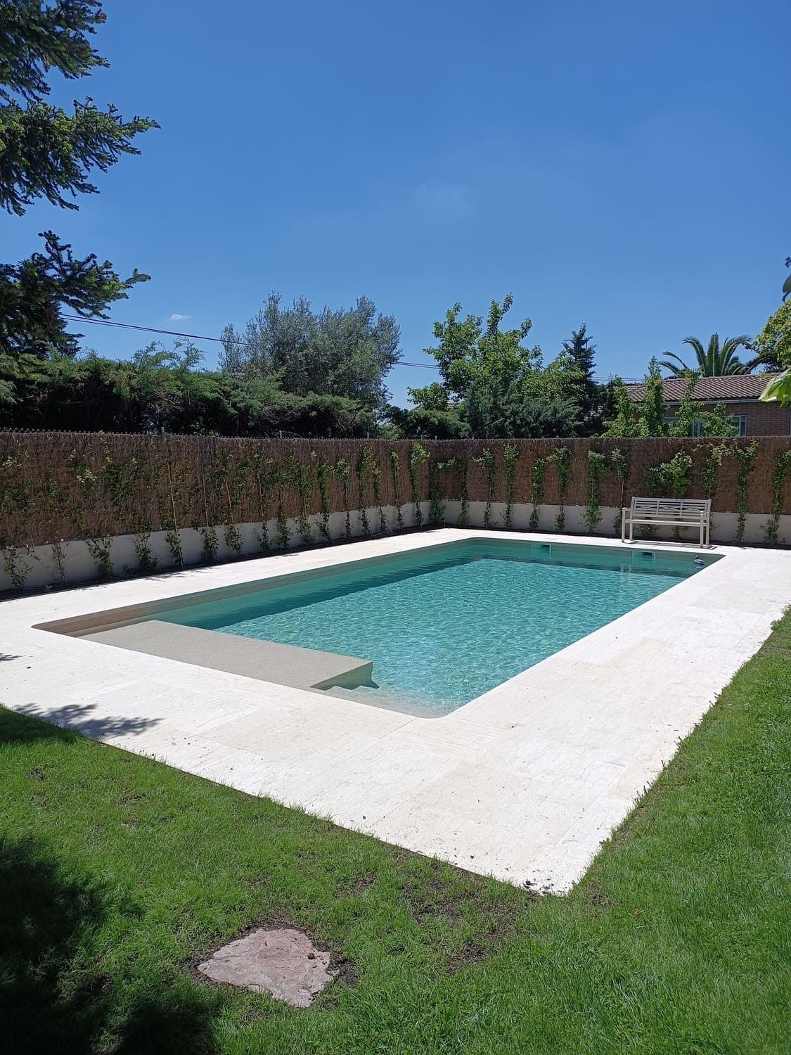 Piscina rectangular rodeada de piedra blanca y césped verde bajo un cielo azul.