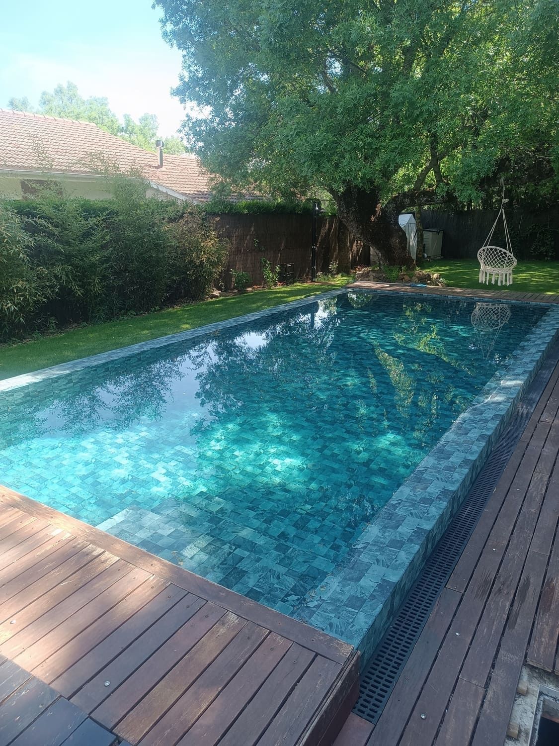 Piscina rectangular de agua turquesa, rodeada de deck de madera y césped, con árboles al fondo.