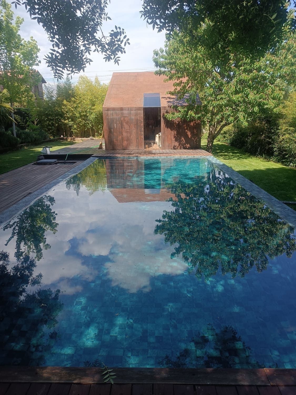 La piscina rectangular refleja el cielo y los árboles, con un pequeño edificio de aspecto rústico al fondo.