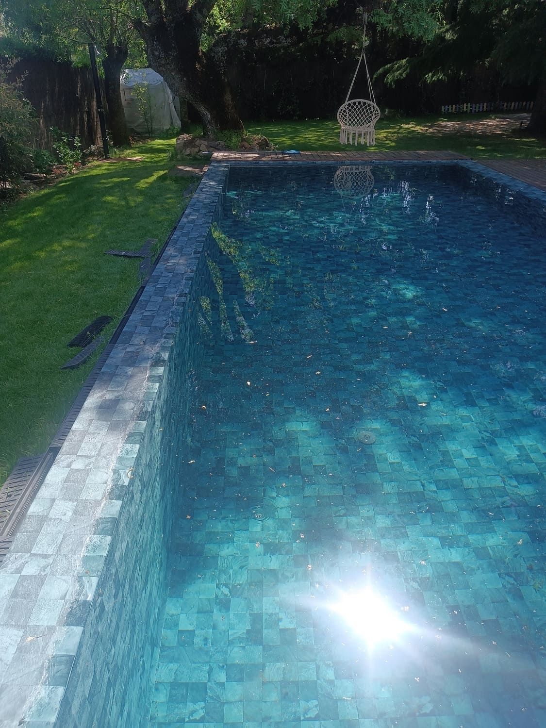 Piscina rectangular de agua turquesa, rodeada de azulejos grises, césped verde y árboles. La luz del sol se refleja en la superficie.