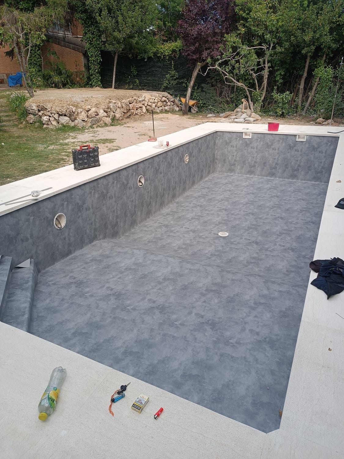 Piscina rectangular vacía con interior gris oscuro y marco de hormigón. Herramientas y árboles visibles al fondo.