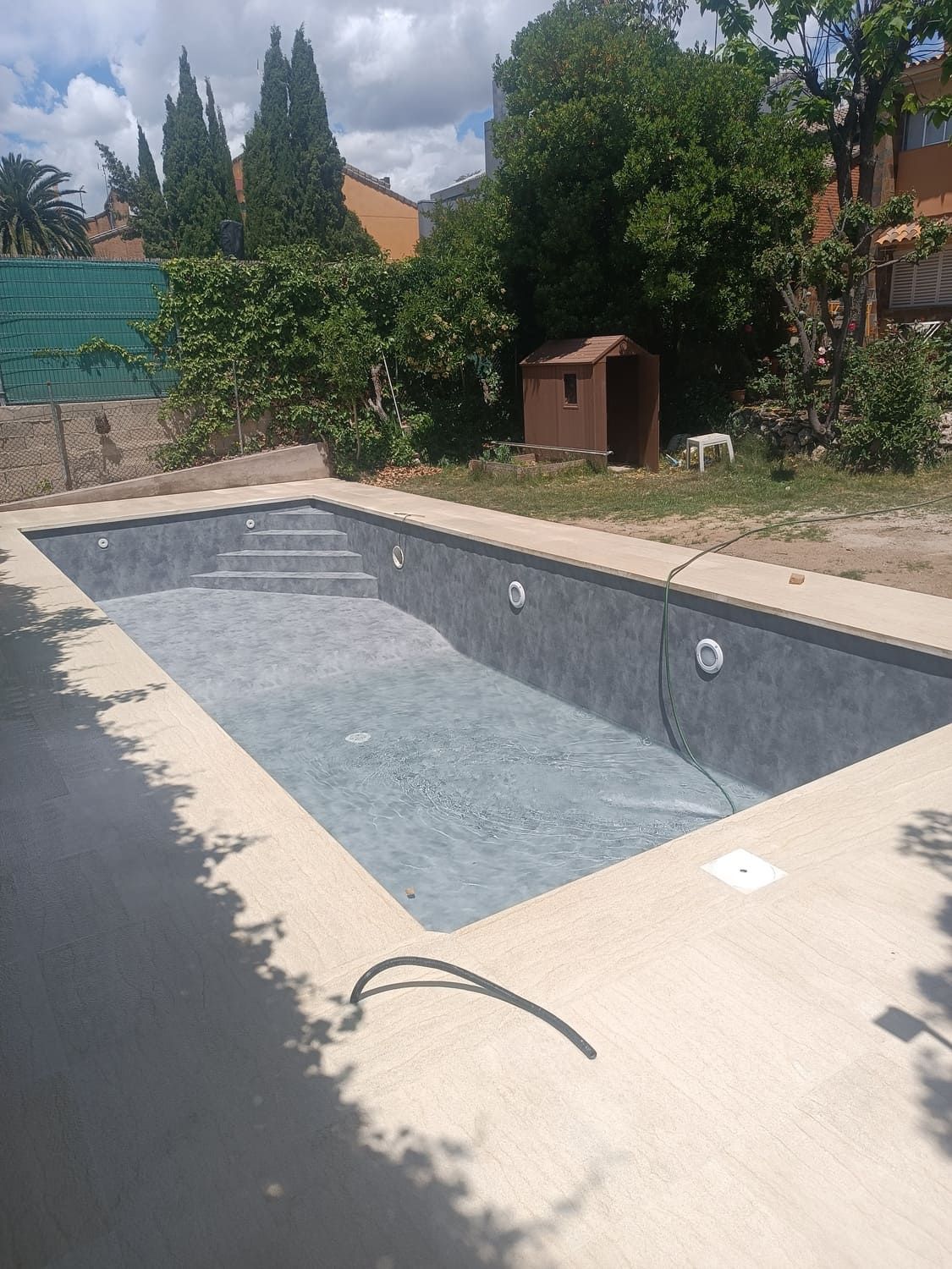 Piscina rectangular vacía con borde de hormigón. Arbustos y árboles verdes al fondo.