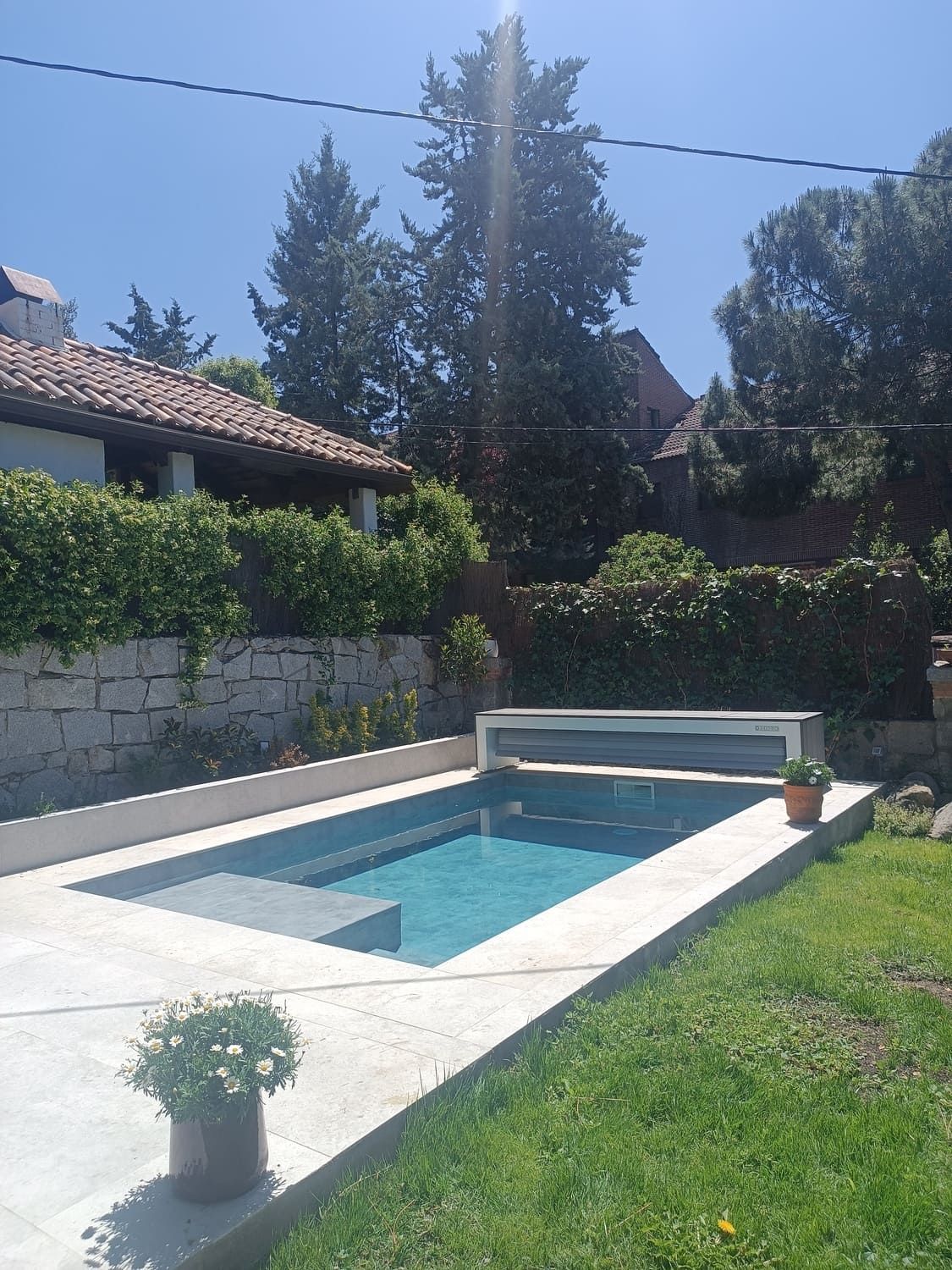 Piscina rectangular de agua azul y cubierta blanca en un patio trasero, rodeada de césped y vegetación.