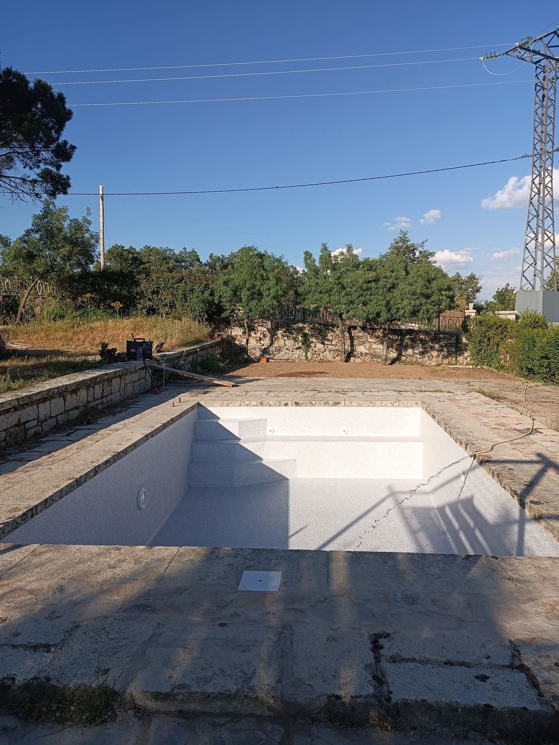 Piscina rectangular vacía, entorno de hormigón, cielo azul, árboles al fondo.