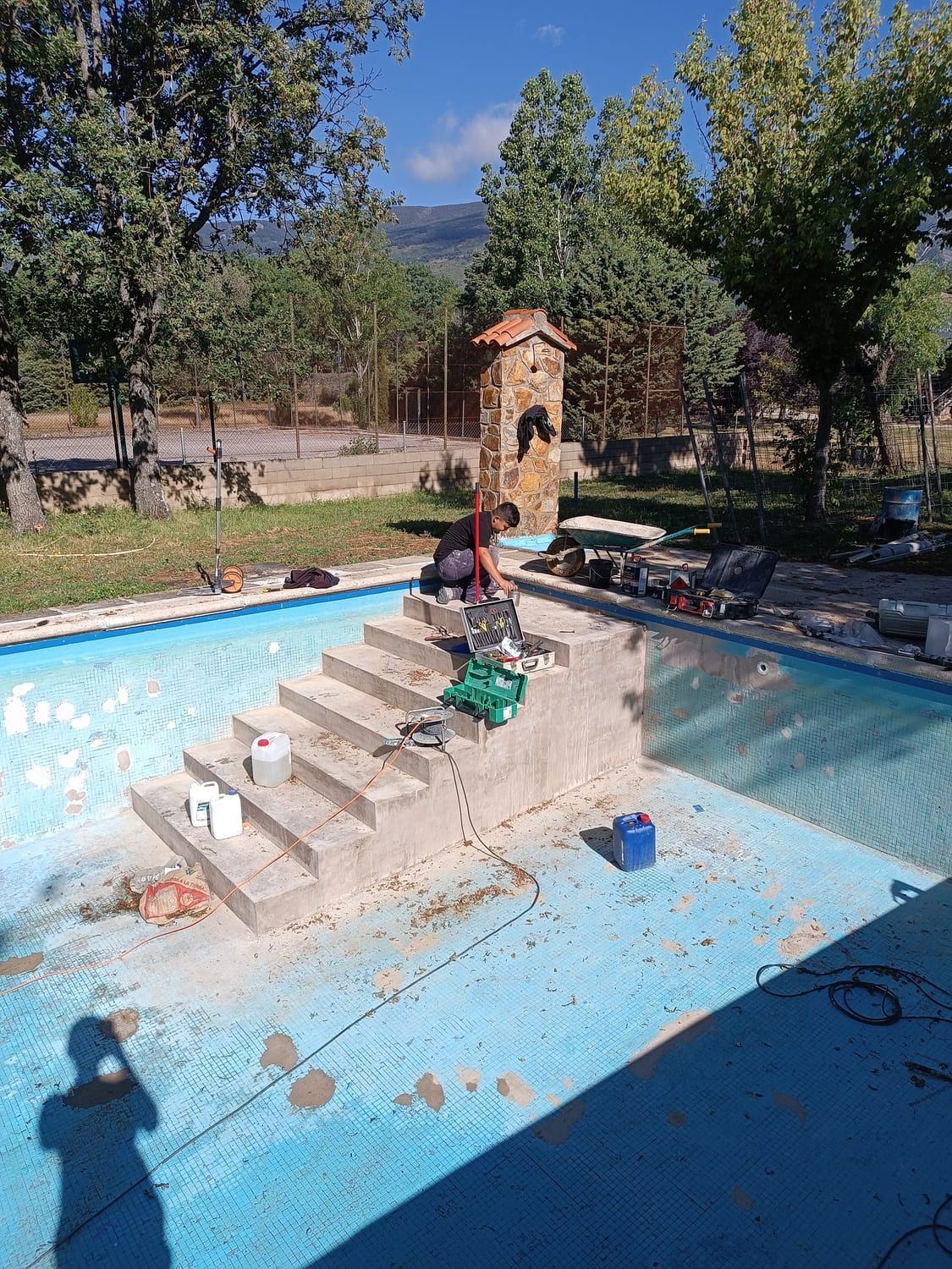 Piscina con escaleras en construcción; herramientas y suministros en las escaleras. Pequeña estructura de piedra al fondo, árboles y cielo azul.