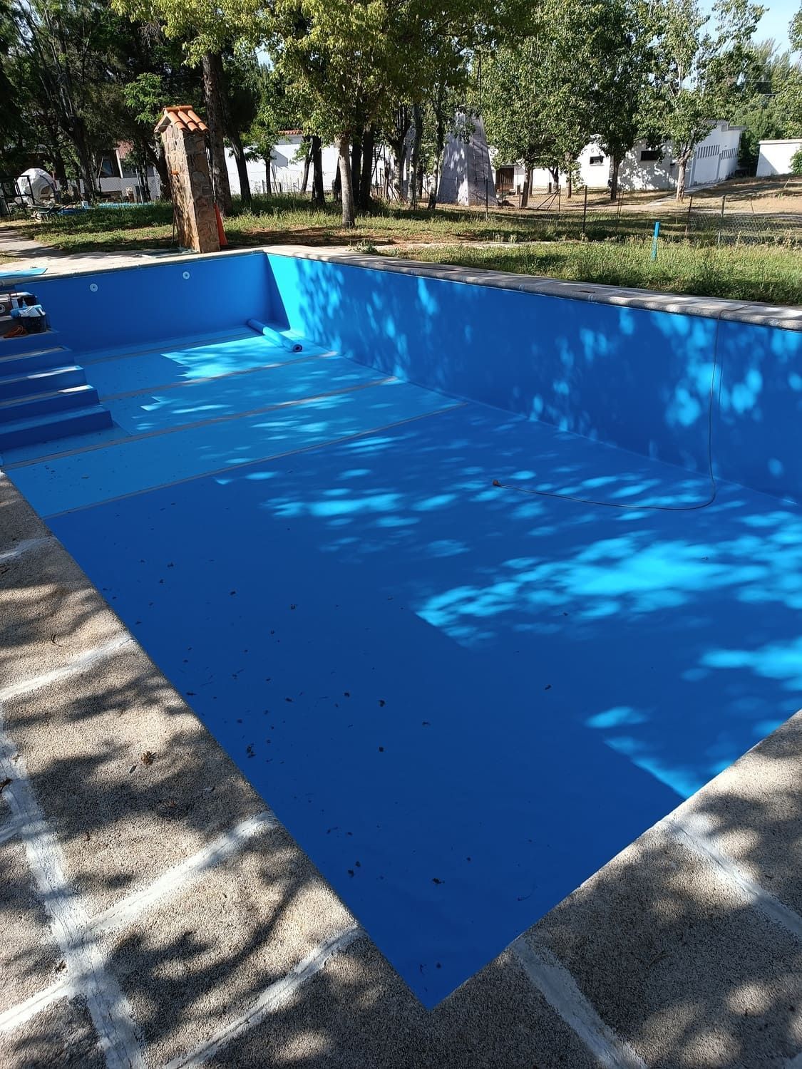 Piscina azul vacía al aire libre, rodeada de hormigón y árboles.