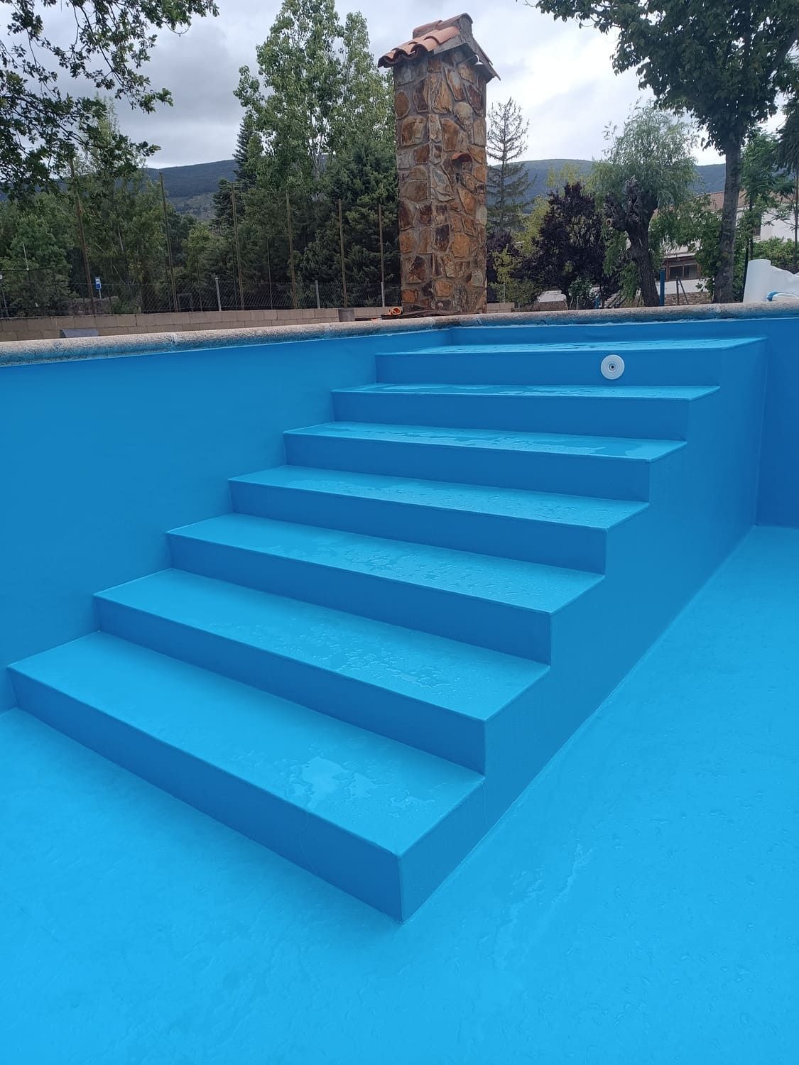 Piscina azul con escalones, entorno al aire libre, pilar de piedra al fondo.
