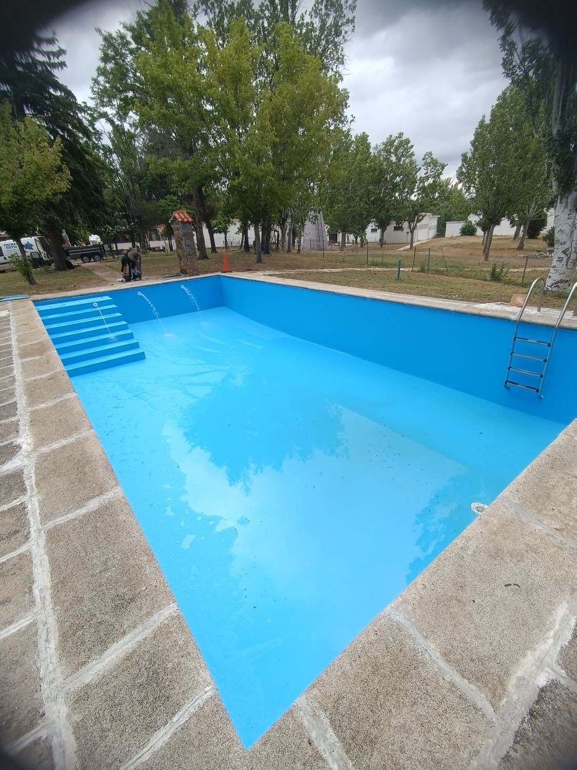 Piscina azul vacía con escaleras y escalerilla, bordeada de piedra y situada en una zona de césped con árboles.