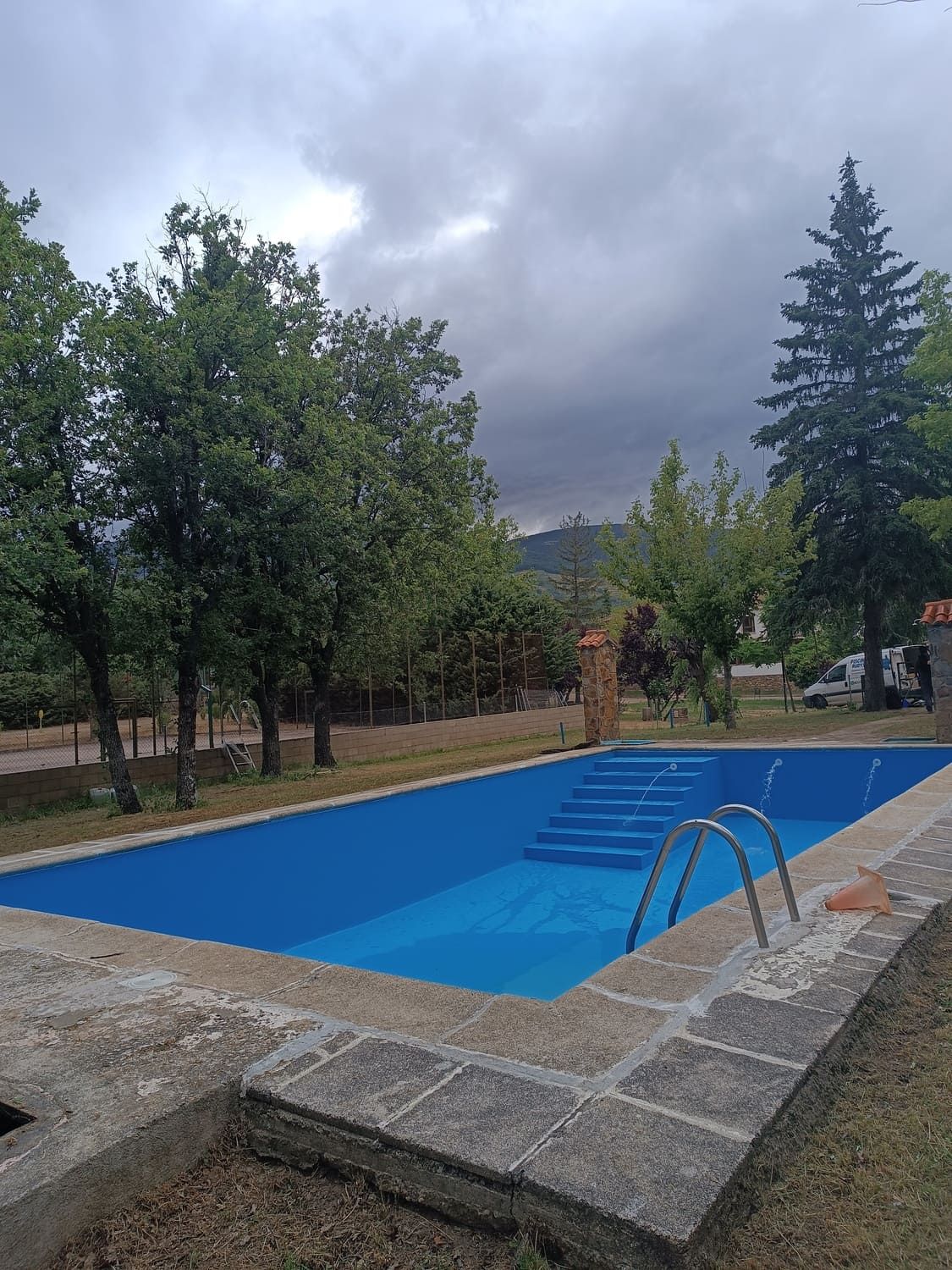 Piscina exterior rectangular de agua azul. Se ven árboles, un cielo nublado y un borde de piedra.