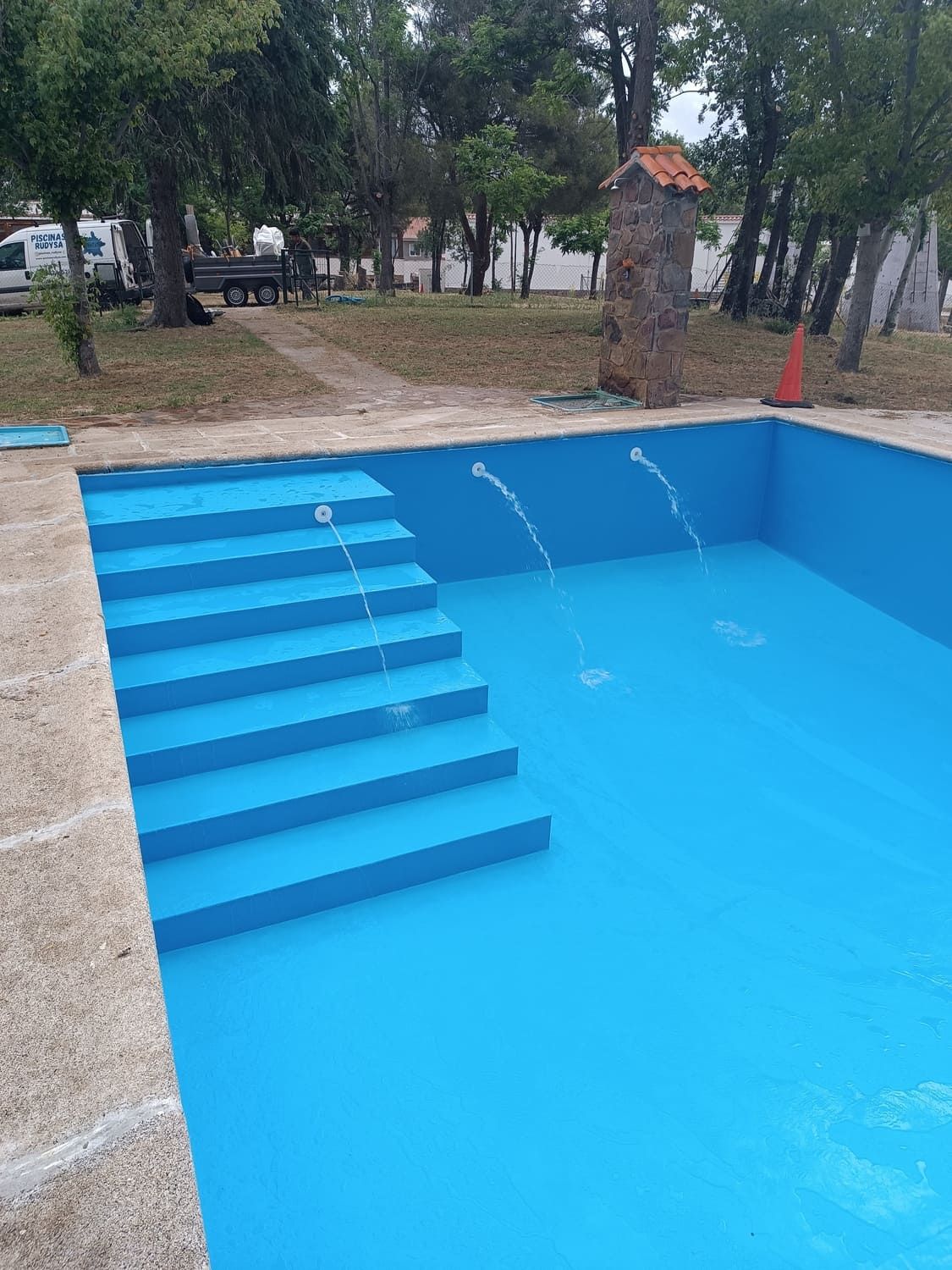 Piscina vacía pintada de azul con escaleras y fuentes. Zona exterior con árboles y parque.