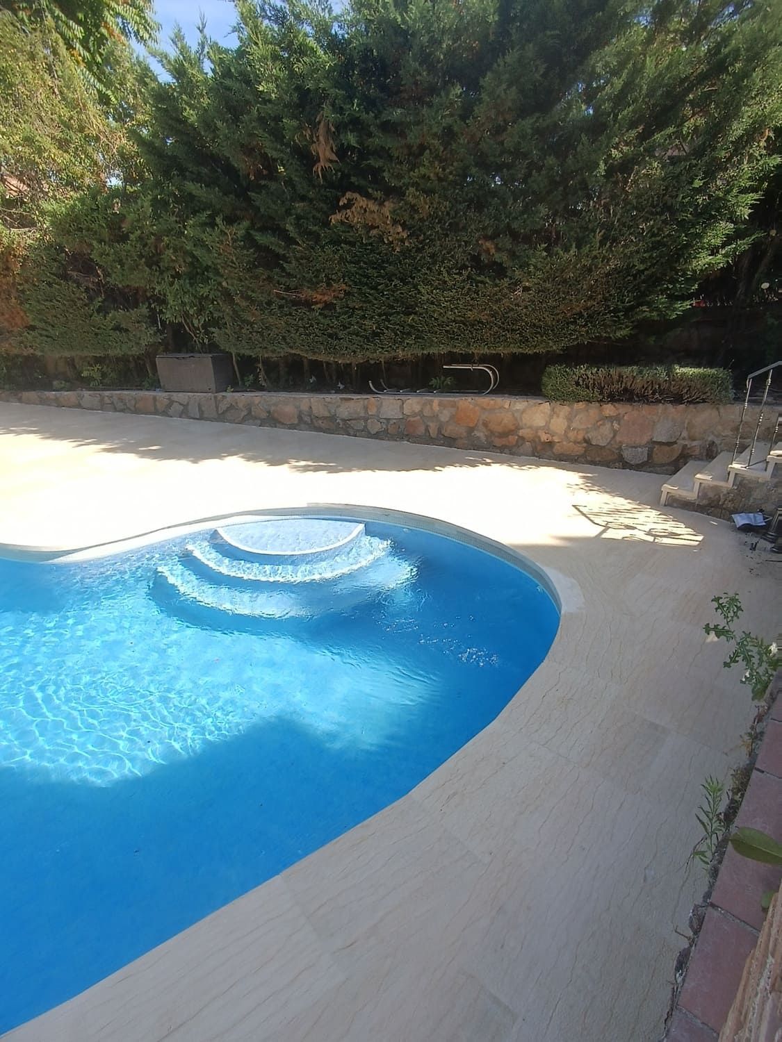 Piscina con agua azul y escaleras, rodeada de hormigón beige y vegetación.