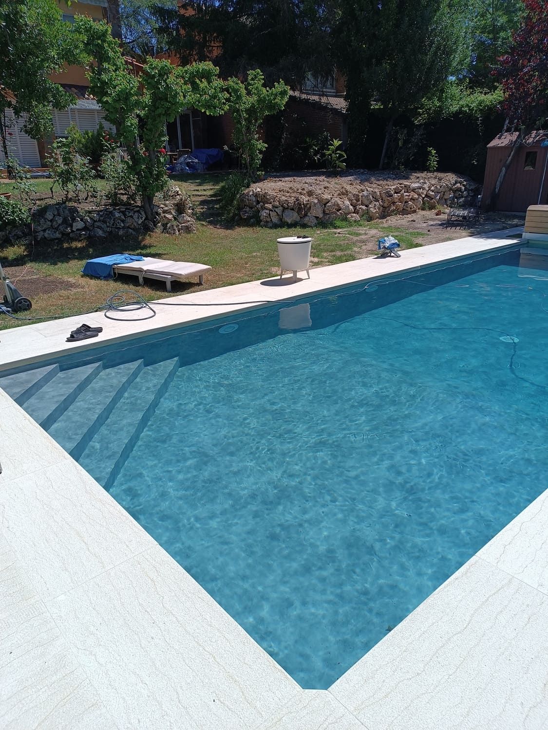 Piscina al aire libre con agua azul y borde de hormigón blanco. Césped verde y árboles al fondo.