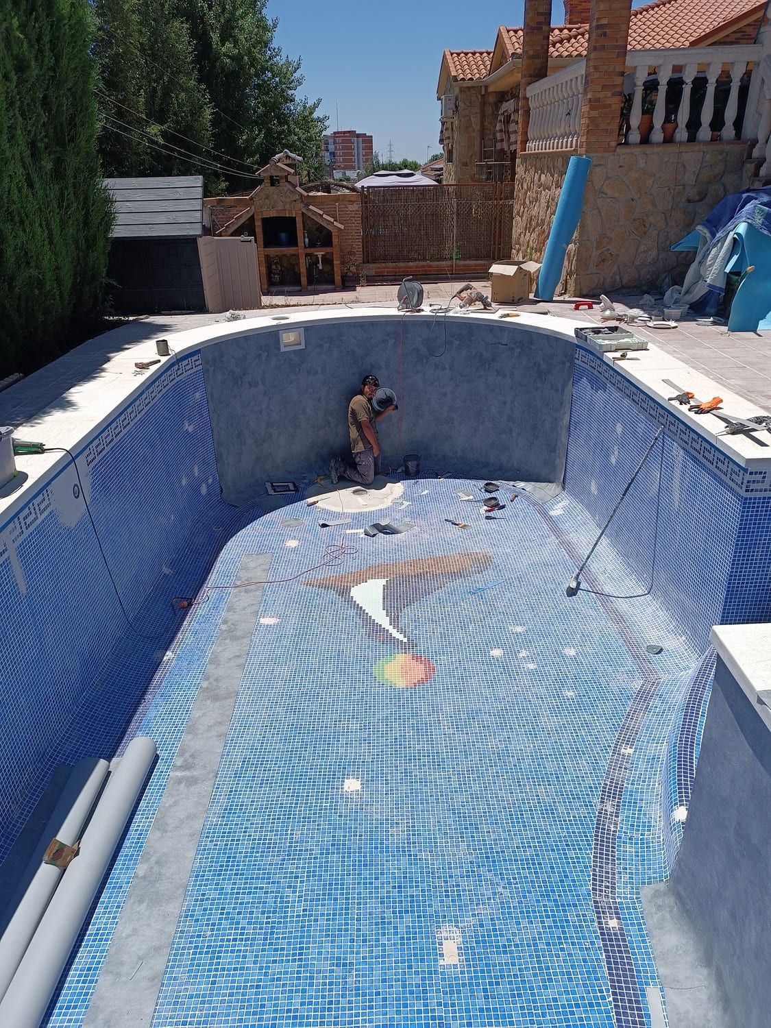 Piscina en construcción, revestida con mosaico azul. Un trabajador está dentro, con herramientas cerca.