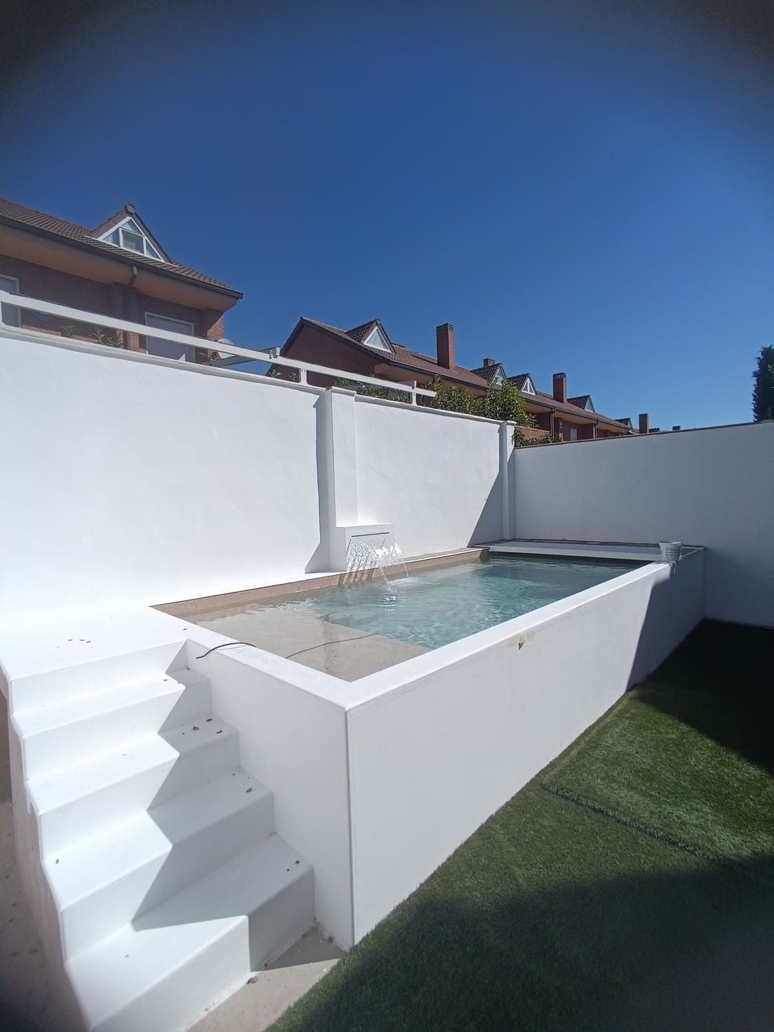 Piscina rectangular de paredes blancas con escalones en un patio trasero, césped verde artificial y cielo azul brillante.