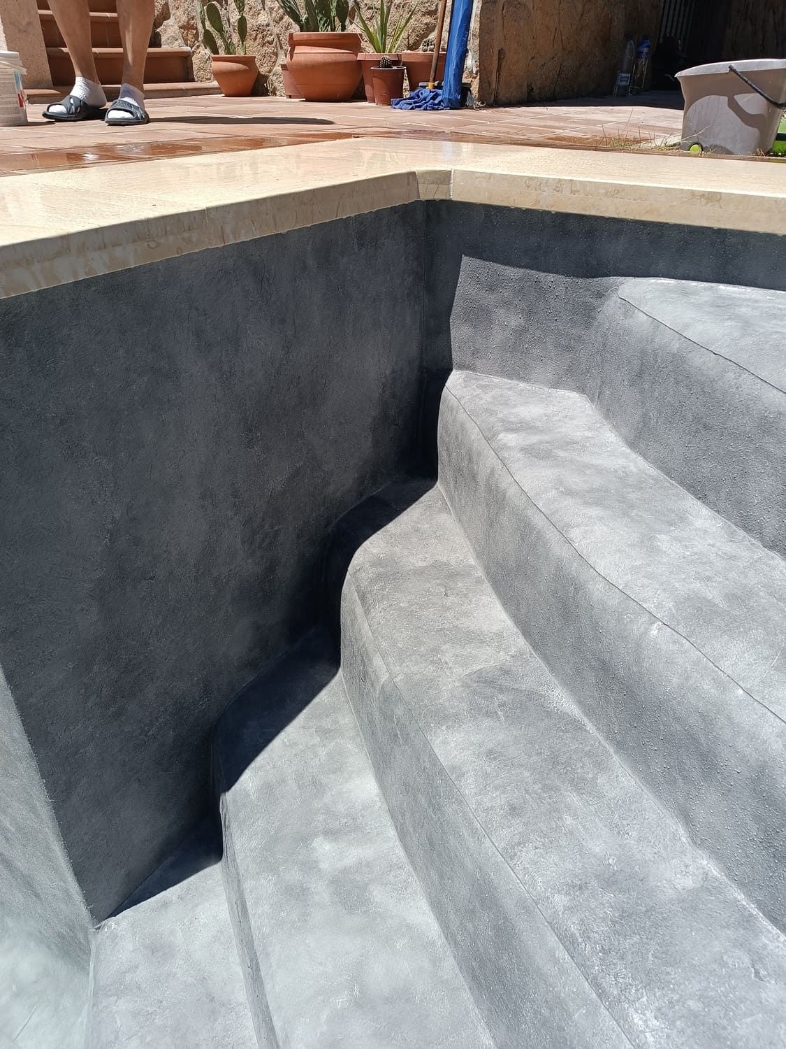 Piscina de paredes grises con escalones. Persona de pie en el borde. Luz solar, exterior.