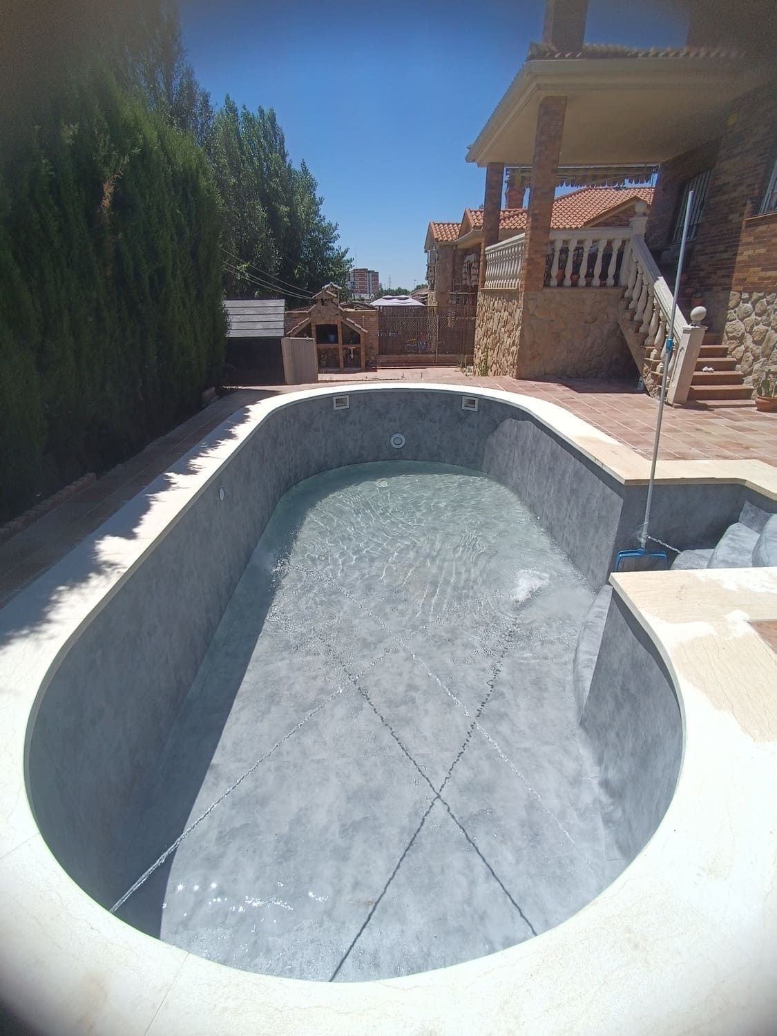 Piscina ovalada llena de agua, rodeada de hormigón. Patio trasero con edificación y árboles.