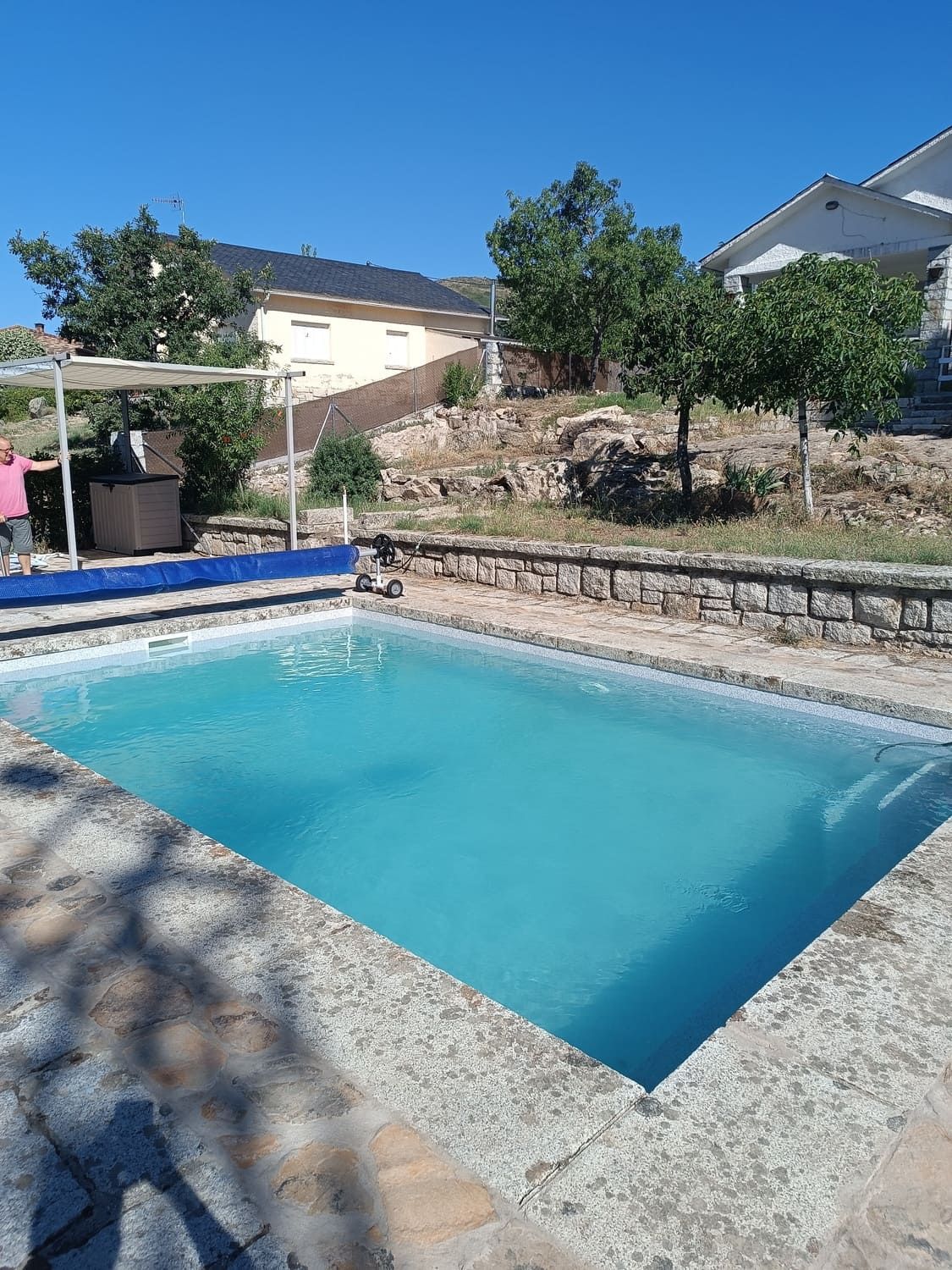 Piscina rectangular con agua azul cristalina, borde de piedra y vegetación circundante bajo un cielo brillante.