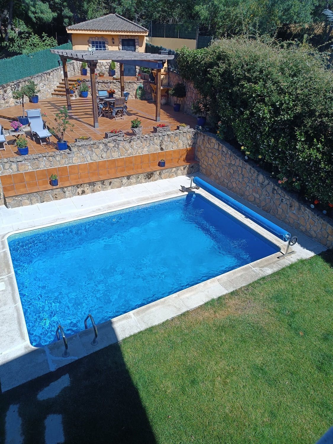 Piscina de agua azul, rodeada de hormigón y césped. Al fondo, una casa, un patio y un jardín.