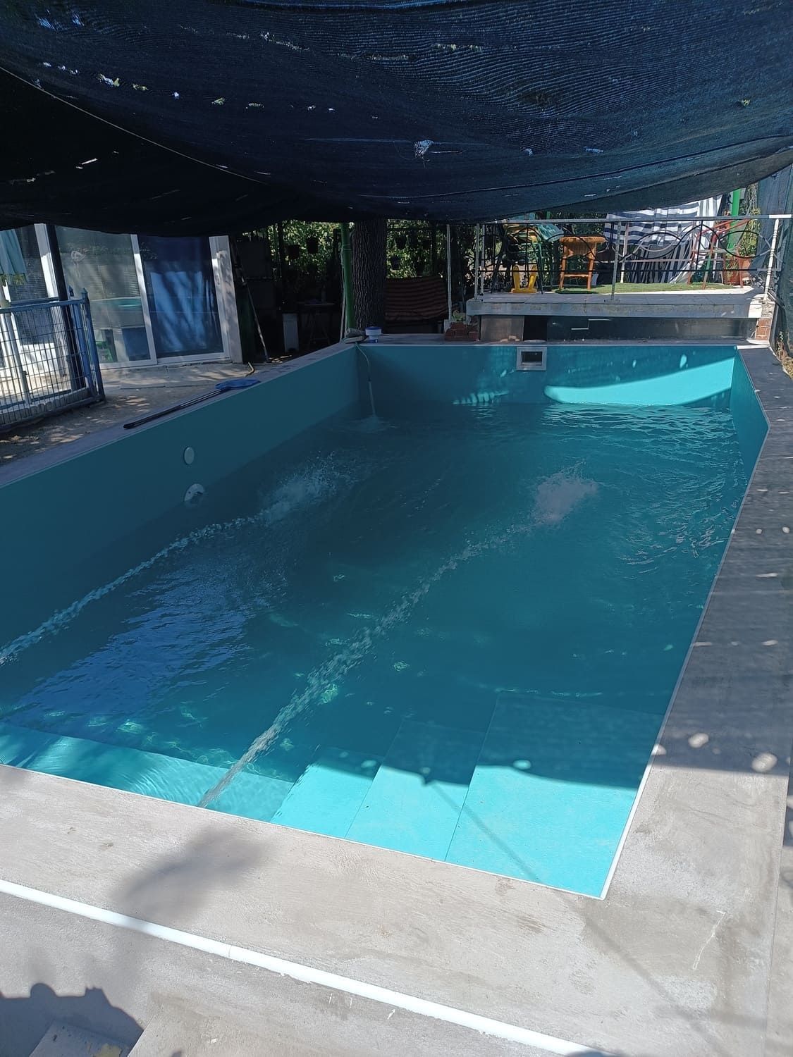 Piscina vacía de color turquesa con escalones, bajo una zona de sombra.