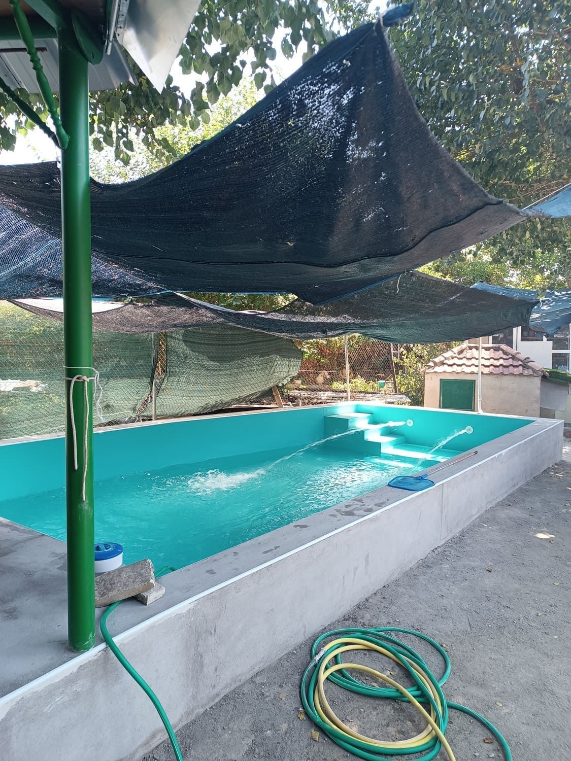 Piscina con agua turquesa bajo un dosel de sombra oscura; tuberías verdes y alrededores de hormigón.