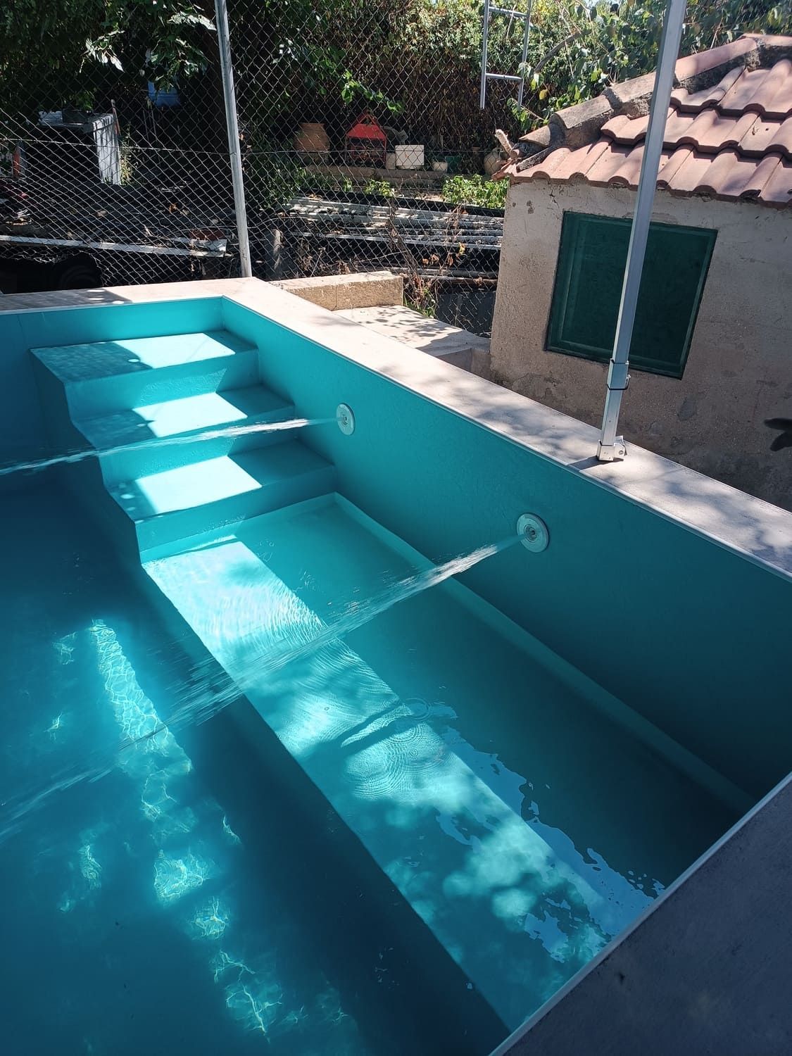 Piscina turquesa con escalones. Agua fluyendo por los chorros. Pequeño edificio con persianas verdes al fondo.