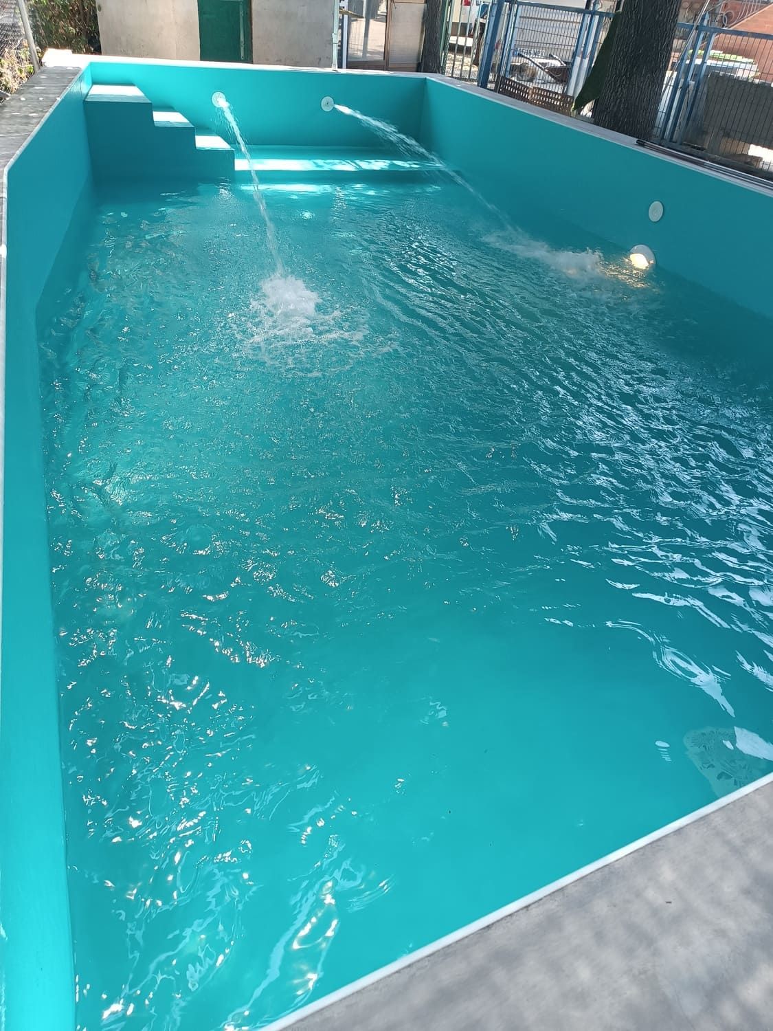 Una piscina turquesa llena de agua. Unas escaleras conducen a la piscina. Chorros de agua salpican en el centro.