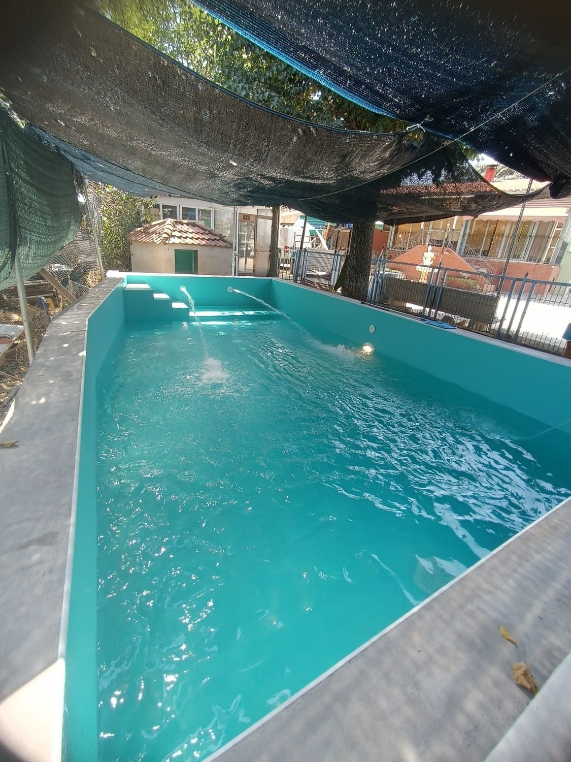 Una piscina al aire libre rectangular llena de agua turquesa, con escalones en un extremo, cubierta por una sombra negra.