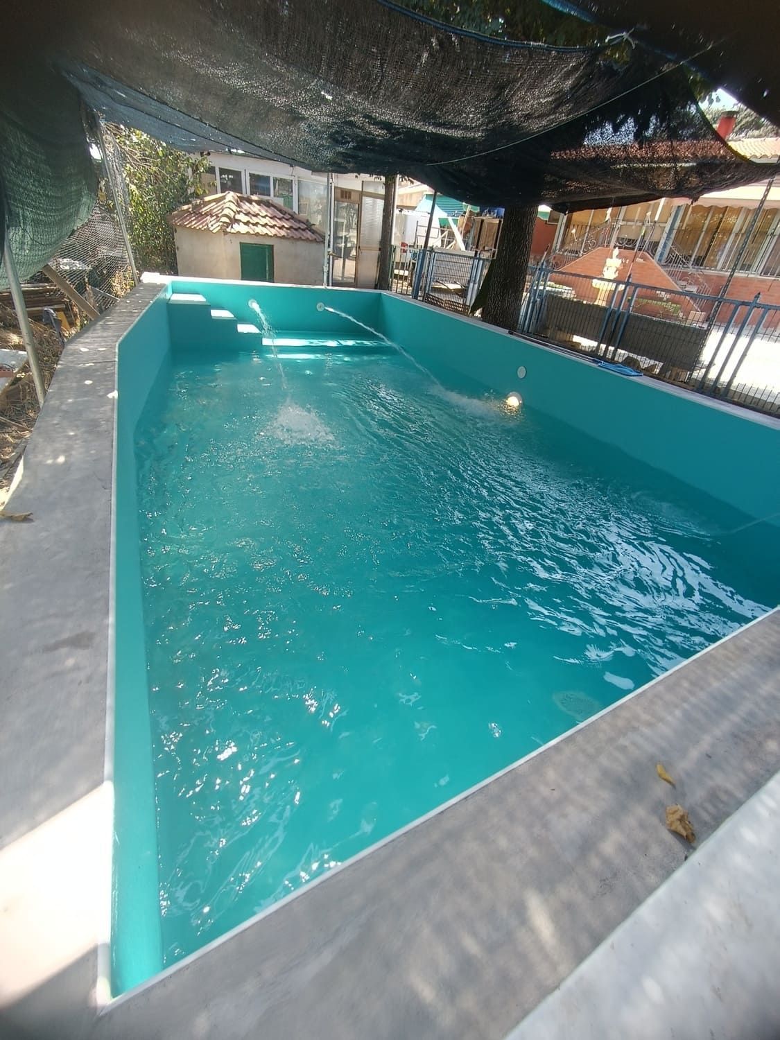 Piscina rectangular de agua turquesa, con escalones en un extremo, bajo zona de sombra.