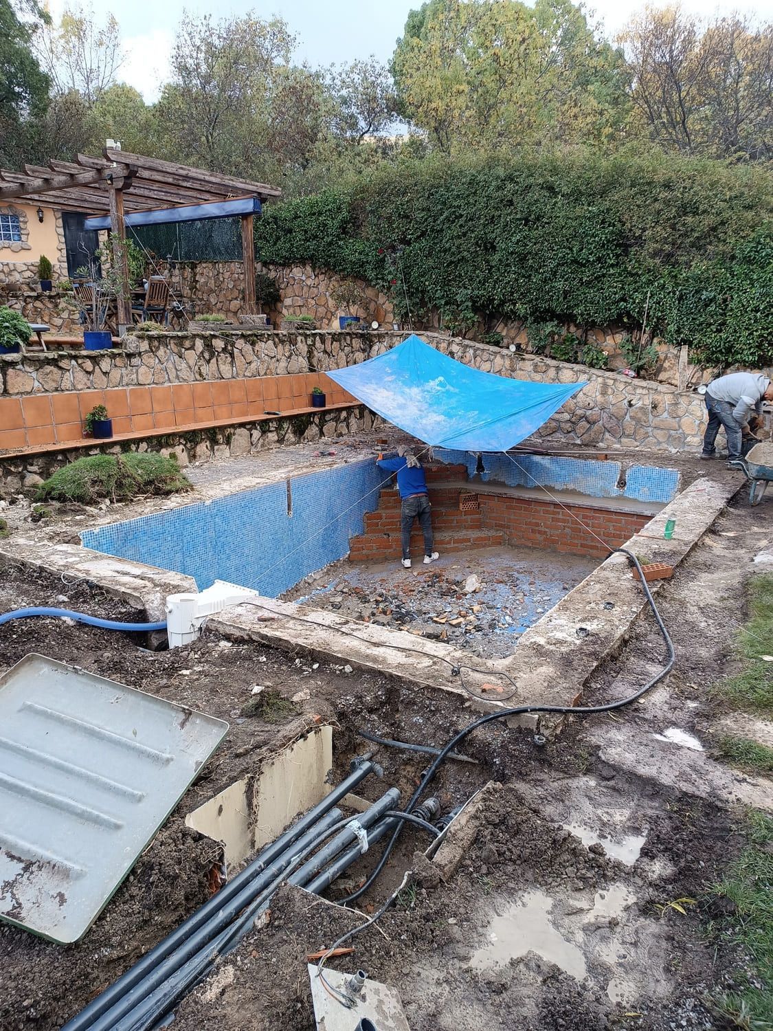 Piscina en remodelación; interior de azulejos azules, bordes de ladrillo, escombros, lona, ​​trabajador, tuberías.