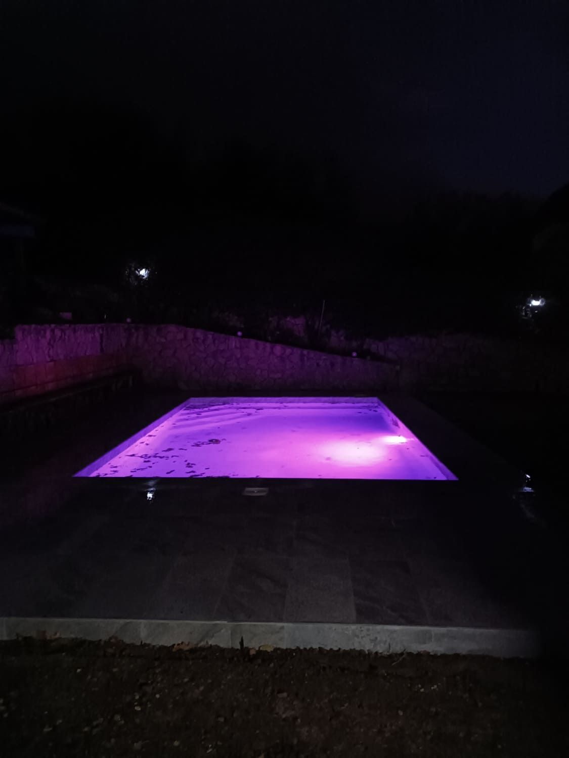 Una piscina rectangular brilla de color púrpura por la noche. El área circundante está oscura.