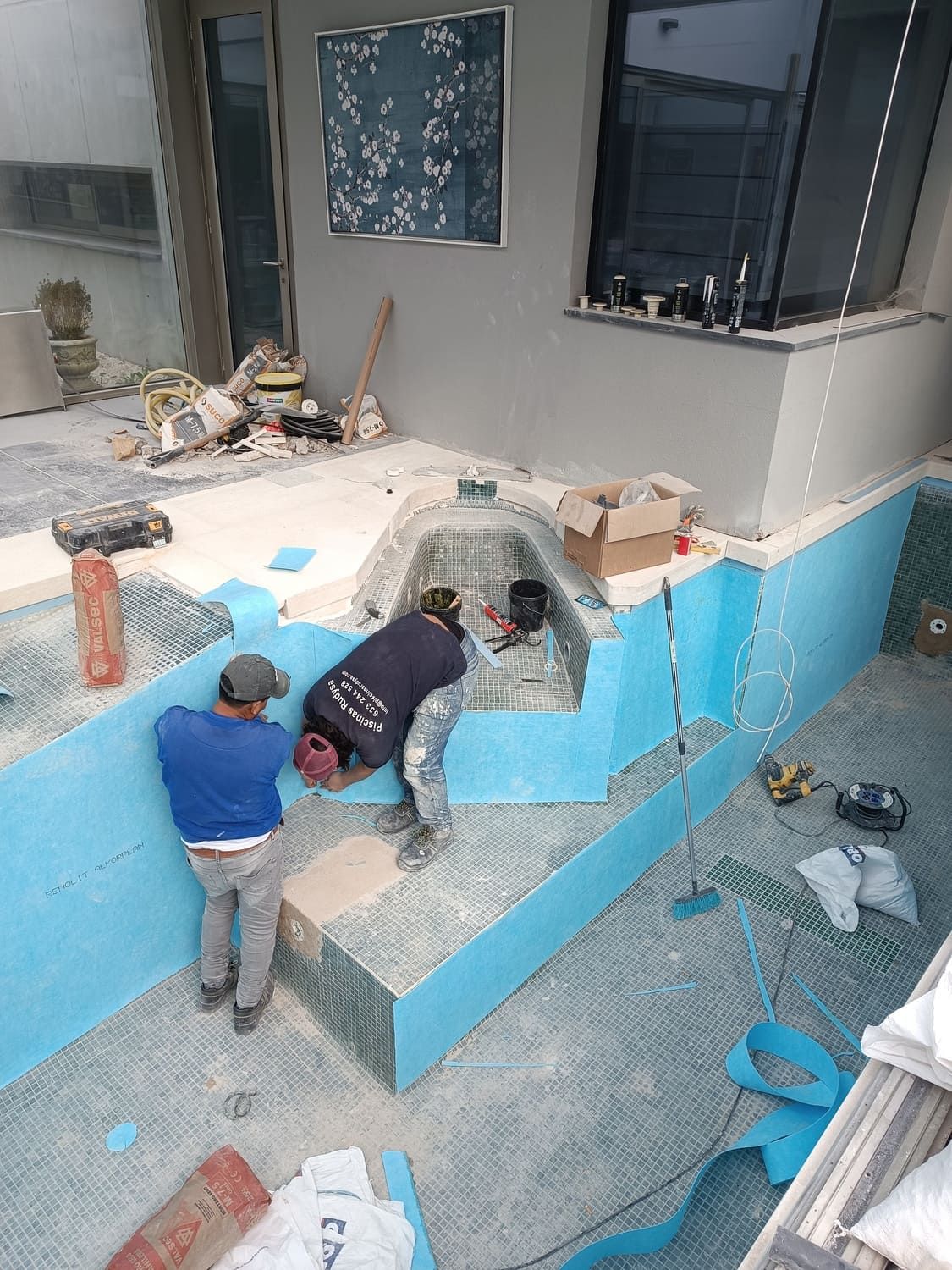 Dos trabajadores alicatando una piscina, azulejos azules, entorno al aire libre.