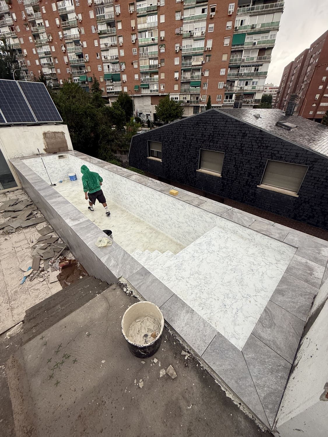 Hombre con sudadera verde en la azotea, instalando baldosas alrededor de una superficie rectangular de color claro; edificios en el fondo.
