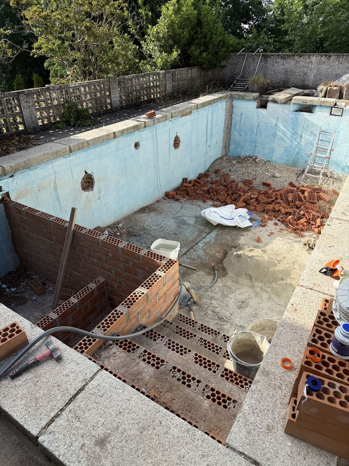 Piscina en construcción; ladrillo, paredes azules, escombros y escalera. Exterior con árboles y pared gris.