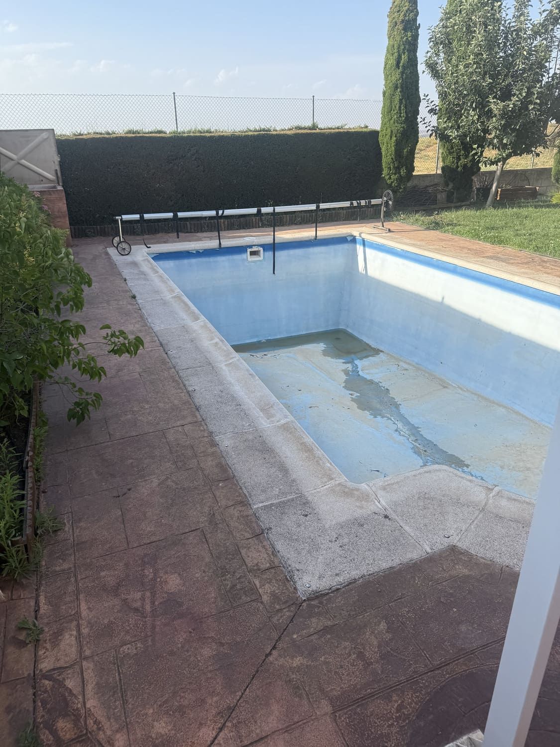 Piscina rectangular vacía con bordes de hormigón, rodeada de patio marrón y seto verde.