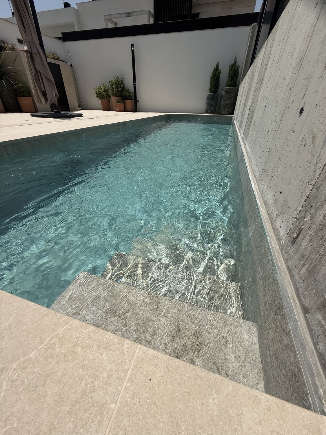 Piscina con escalones incorporados, agua azul claro, pared de hormigón y marco de azulejos beige.